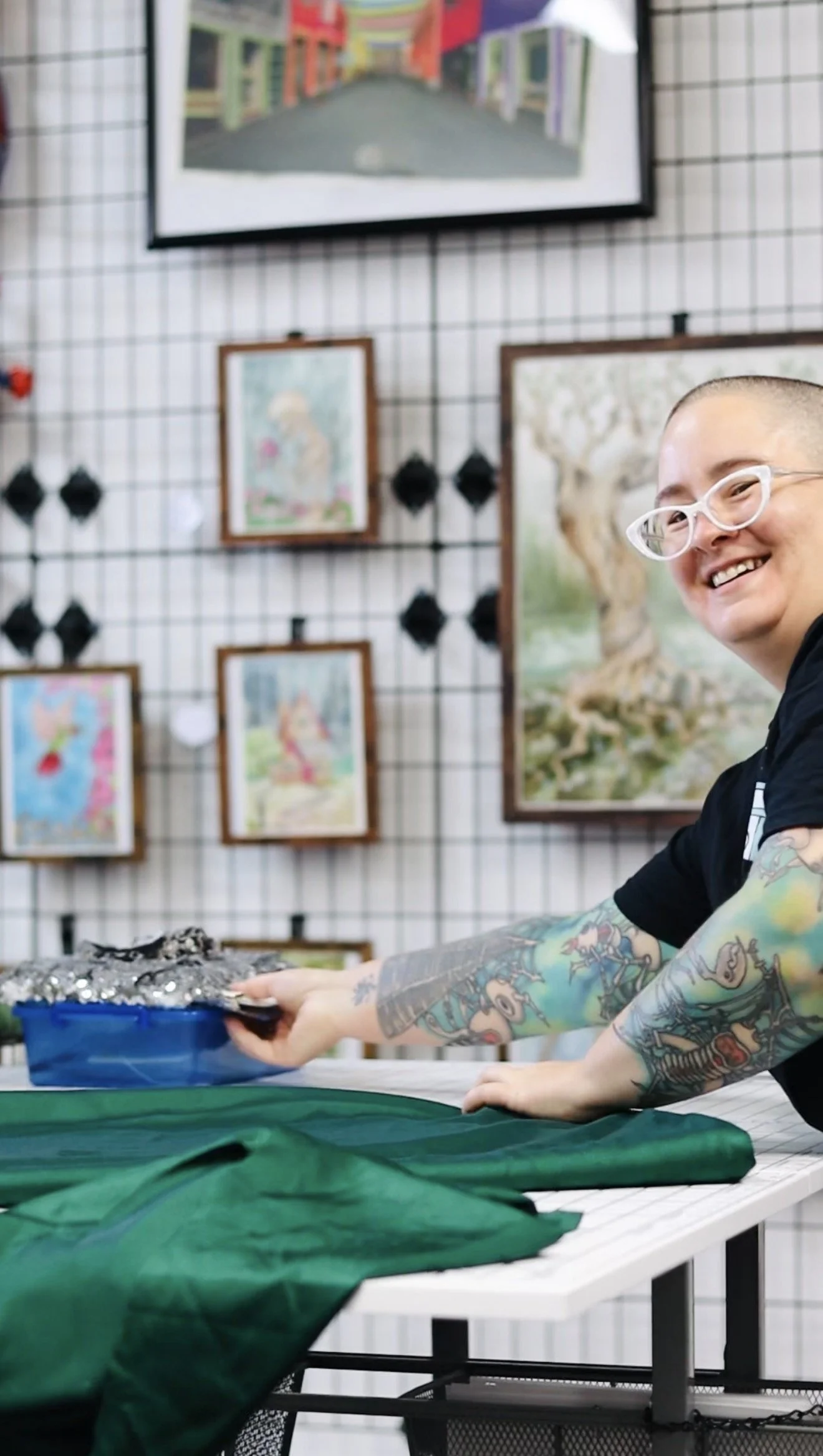 A smiling person with glasses and tattoos on their arms at a fabric cutting table, holding a tray of shiny silver trim, with colorful artwork on a grid wall behind them.