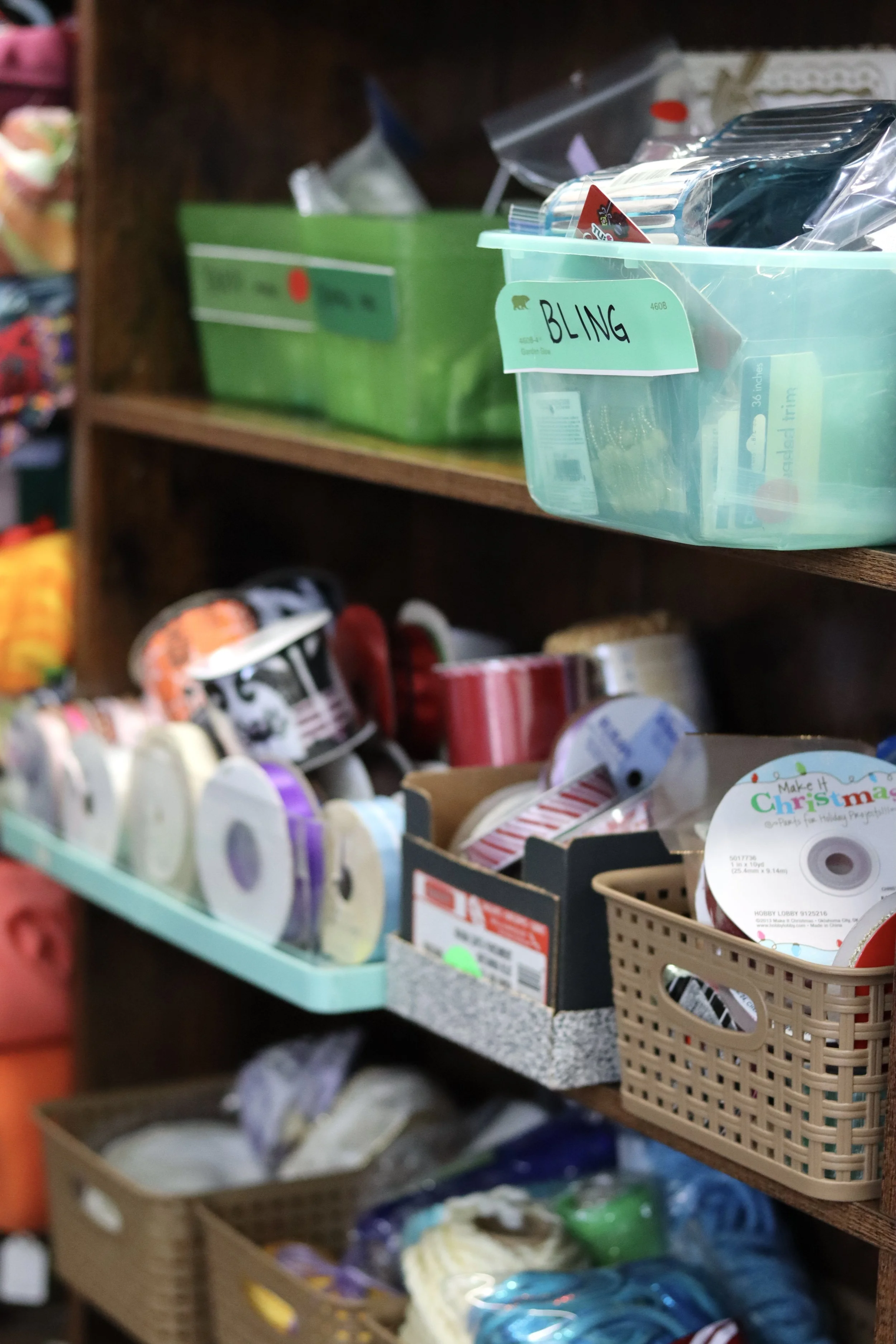 A store shelf with various craft supplies, including spools of ribbon, tape, and decorative items, organized in baskets and boxes.