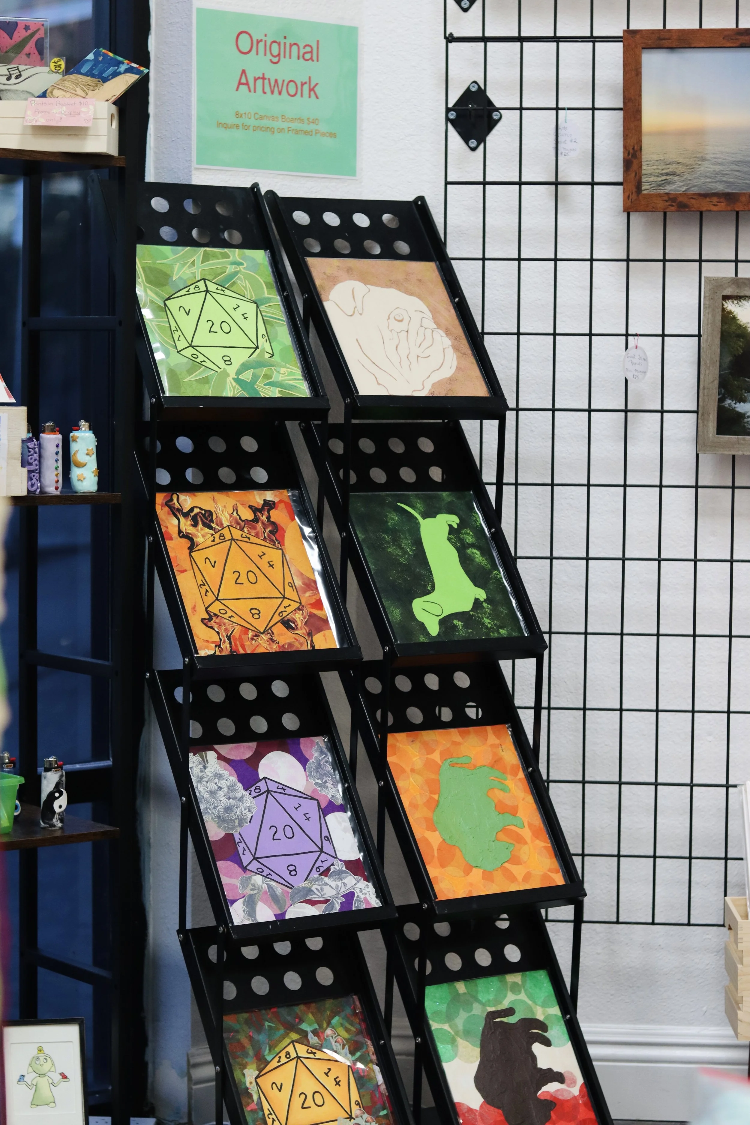 Display of various art pieces on black racks, including colorful paintings of a D20 die, a hand drawing, a green dachshund a purple yorkie, and artistic patterns. Sign on the wall reads 'Original Artwork' with pricing information.