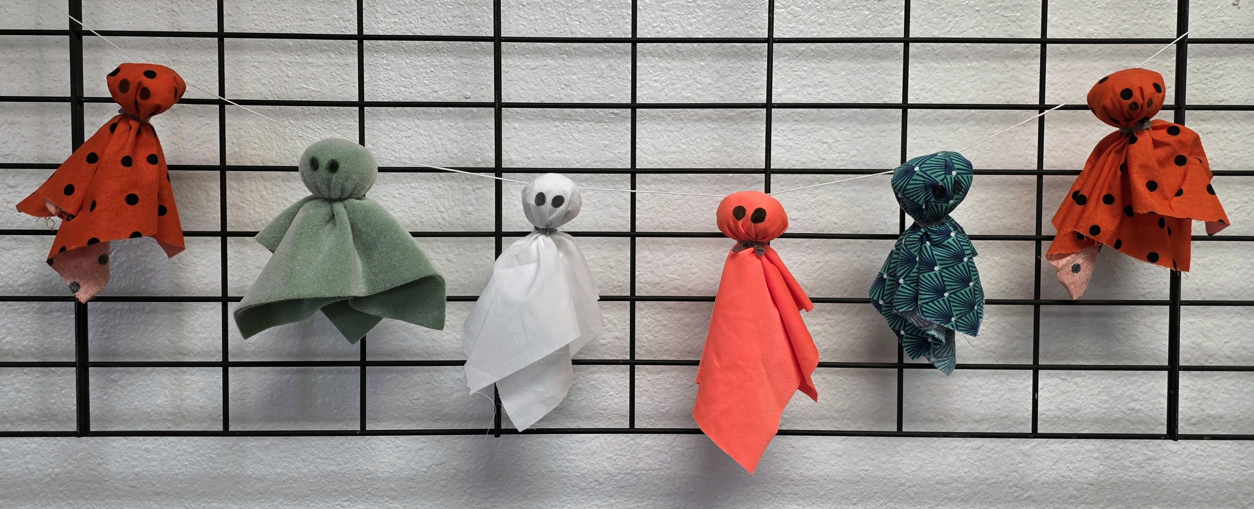 A display of six handmade ghost puppets made from cloth, felt, and string, hanging on a black wire grid against a white wall. Each puppet has a different colored fabric with black details for eyes and mouth.