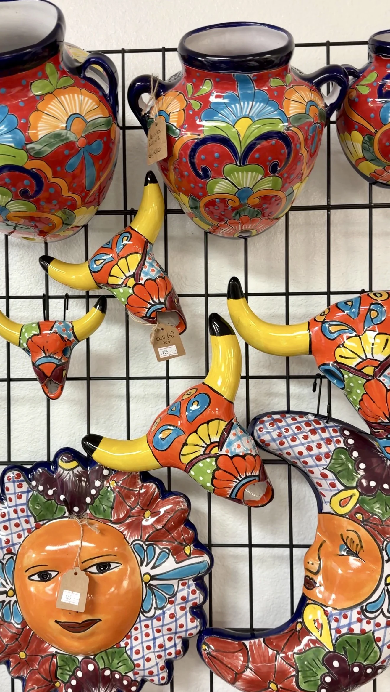 Colorful ceramic Mexican-style vases and wall decorations with floral patterns and painted faces, displayed on a black wire grid.