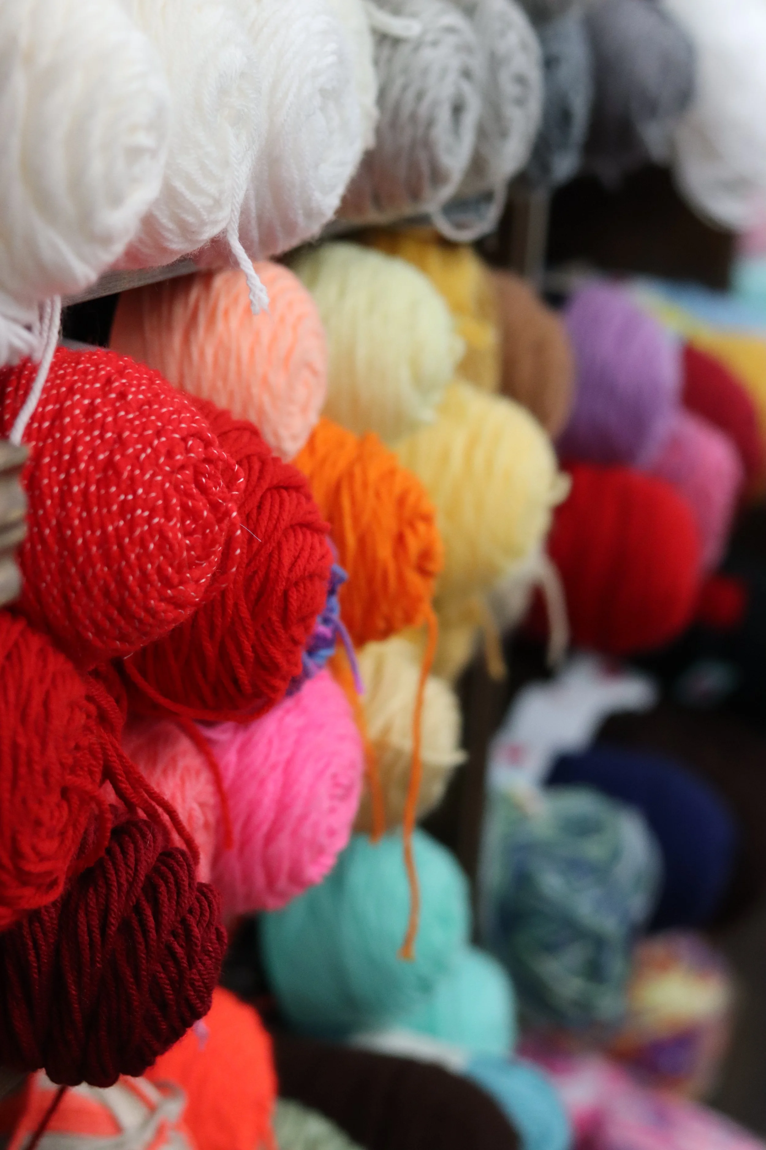 Colorful balls of yarn on shelves, showcasing a variety of colors including red, orange, yellow, purple, teal, pink, brown, gray, cream, and multicolored, in a knitting or craft store.