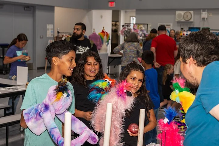Children and adults at a fair or craft event, with children holding colorful sock puppets and chatting with an adult.