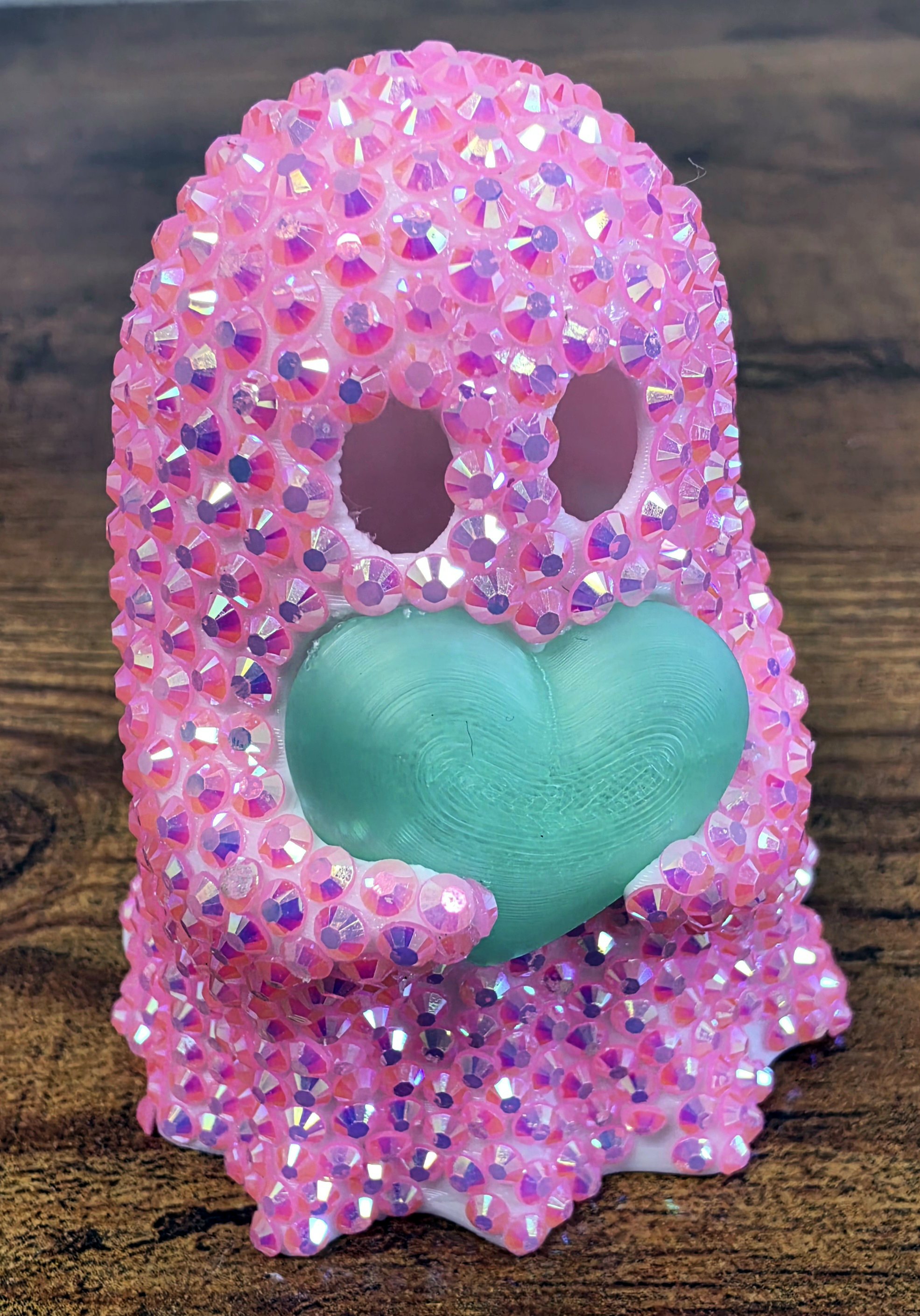 A pink rhinestone decorated 3d printed ghost with a teal-colored heart is placed on a wooden surface.