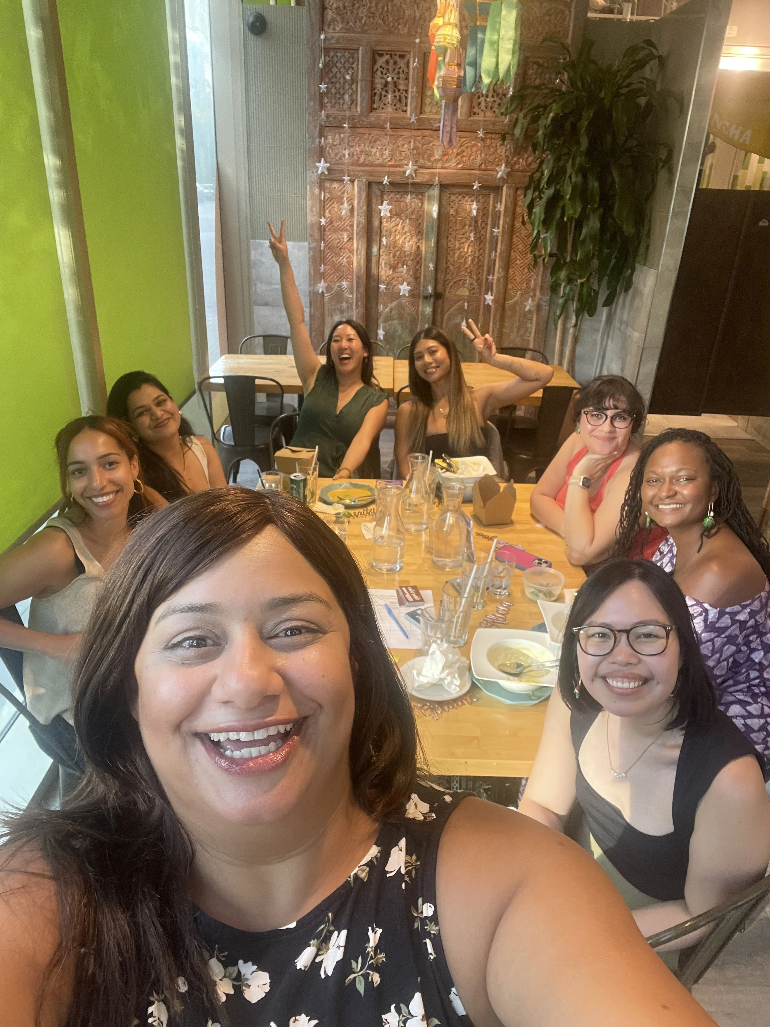 A group of women sitting at a restaurant table, smiling and taking a selfie. The background features a wooden decorative wall, green themed staff uniforms or aprons hanging, and a large potted plant. The table is filled with glasses, bowls, and food containers.