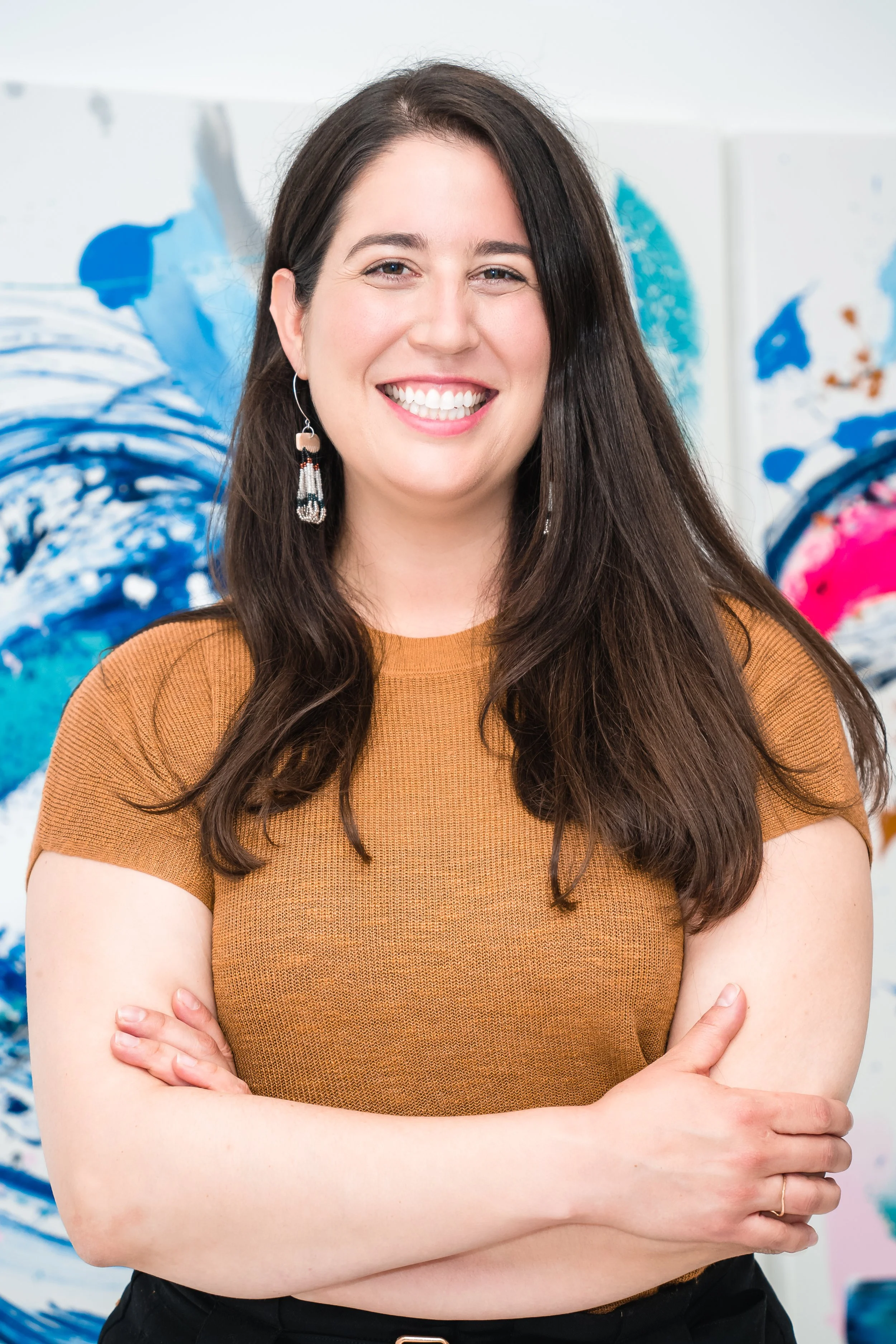 A woman with long dark hair, smiling, wearing a brown short-sleeved top, earrings, standing with arms crossed in front of an abstract blue and pink artwork.