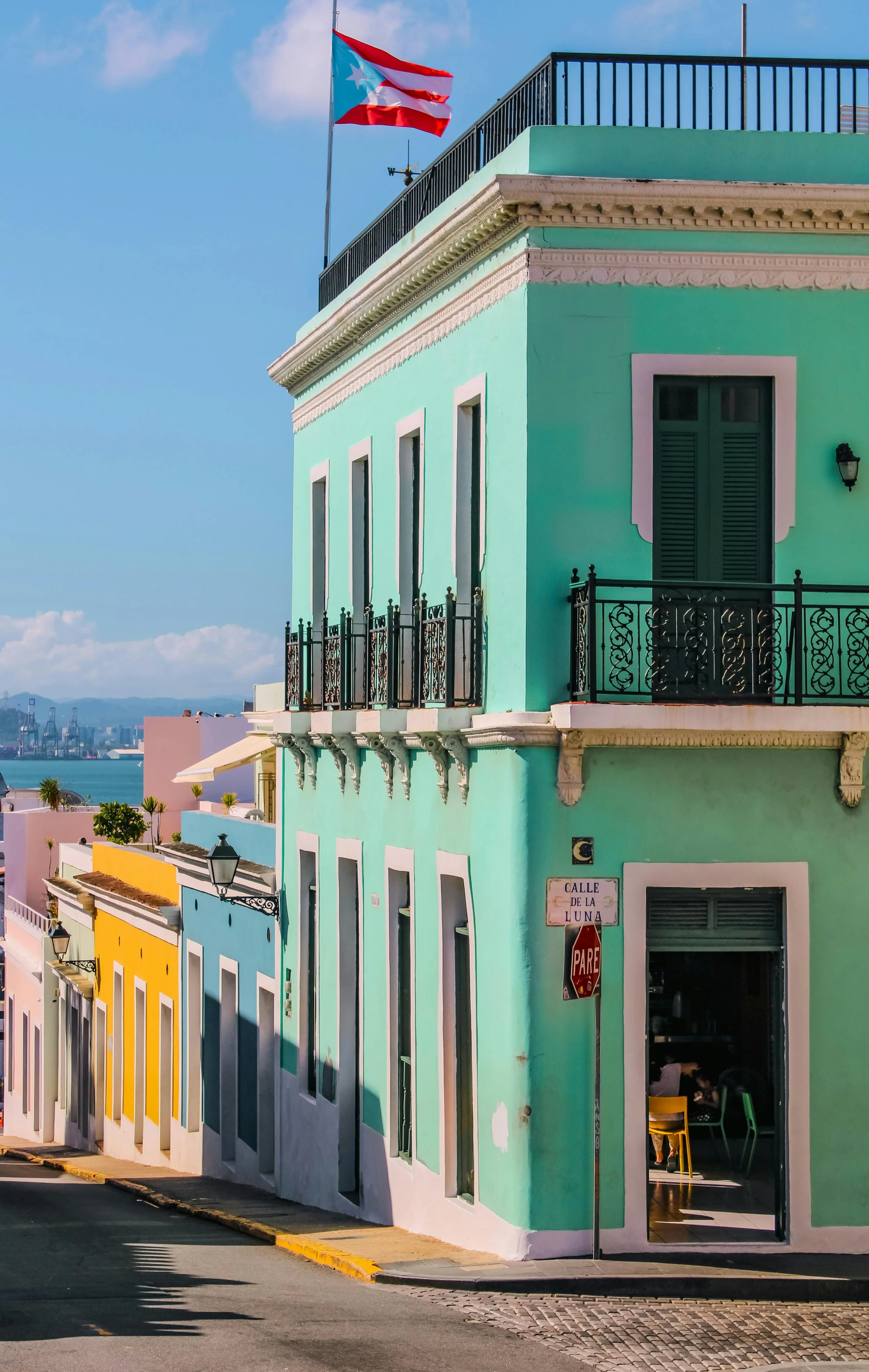 Colorful building on a street corner with pastel green, yellow, blue, and pink facades, featuring wrought iron balconies, open window shutters, and a sign that reads 'Calle de la Luna.' The Puerto Rican flag is flying on top of the building. The street has a yellow curb and a stop sign.