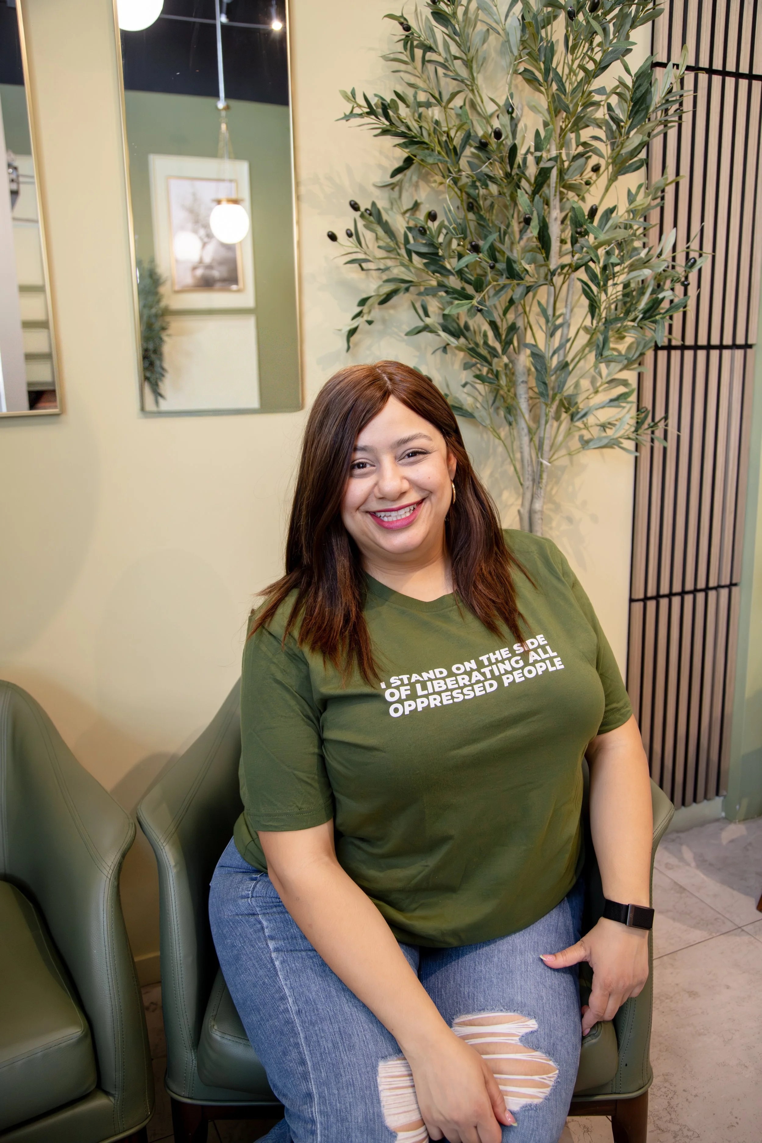 A woman with brown hair, wearing a green T-shirt and ripped jeans, sitting on an olive-green chair, smiling at the camera in an indoor setting with decorative mirrors and a tall plant in the background.