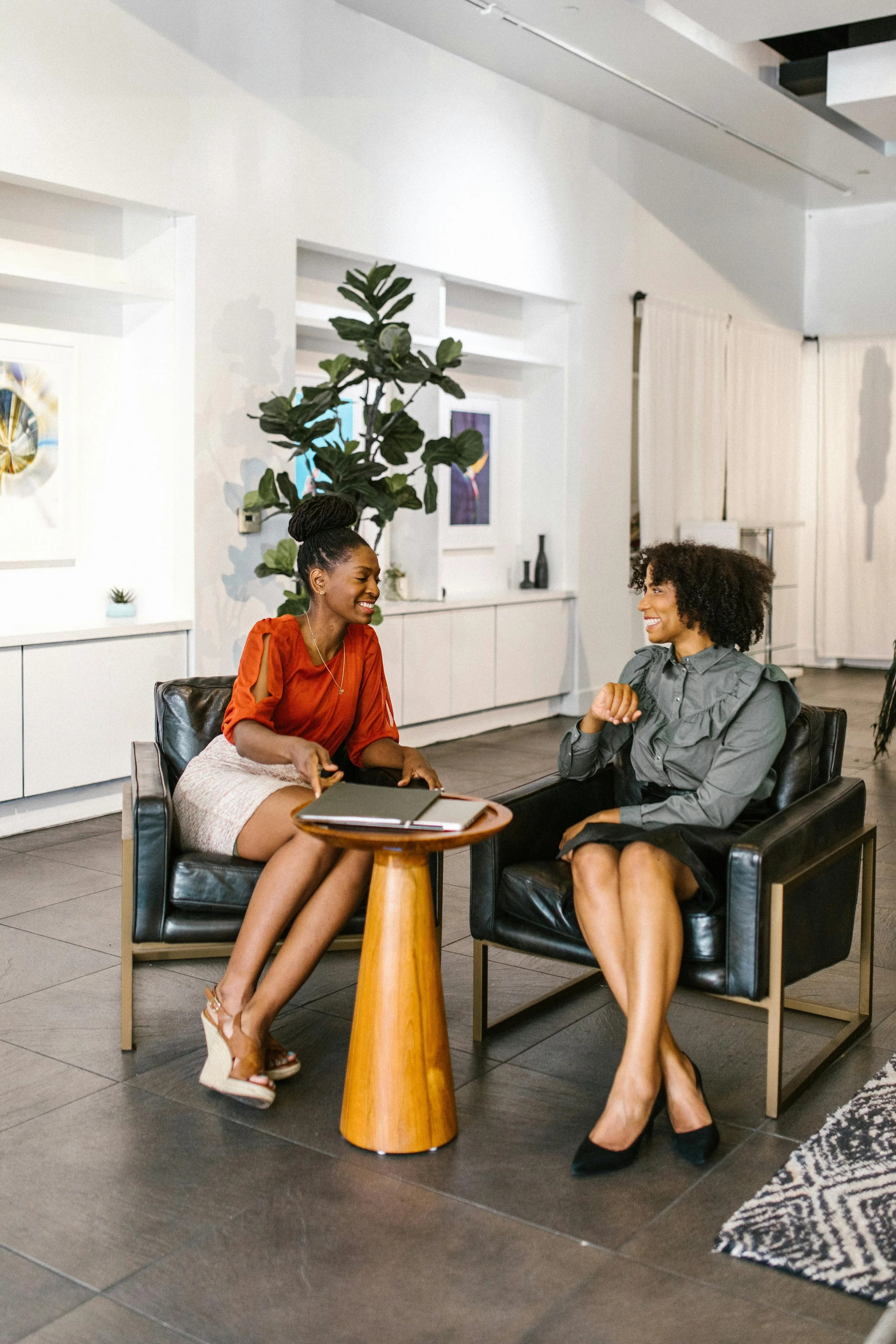 Two women sitting on black leather chairs in a modern indoor space, engaged in conversation, with a small round wooden table between them. The woman on the left is wearing an orange blouse and a light-colored skirt, while the woman on the right is wearing a gray blouse and black shoes. There is a large green plant in the background and artwork on the white walls.