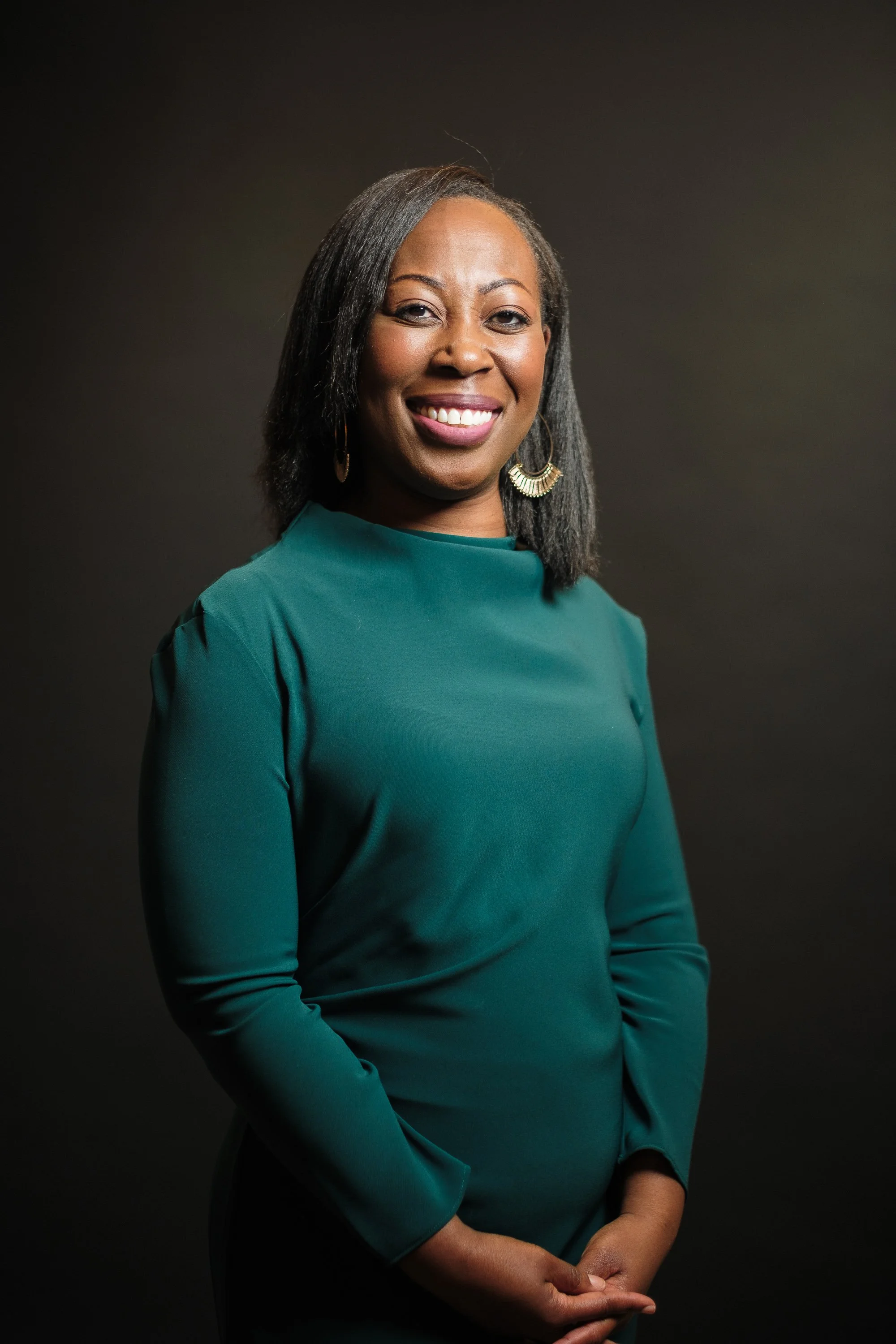 A smiling African American woman with shoulder-length straight hair wearing a teal long-sleeve top, gold earrings, posing against a plain dark background.