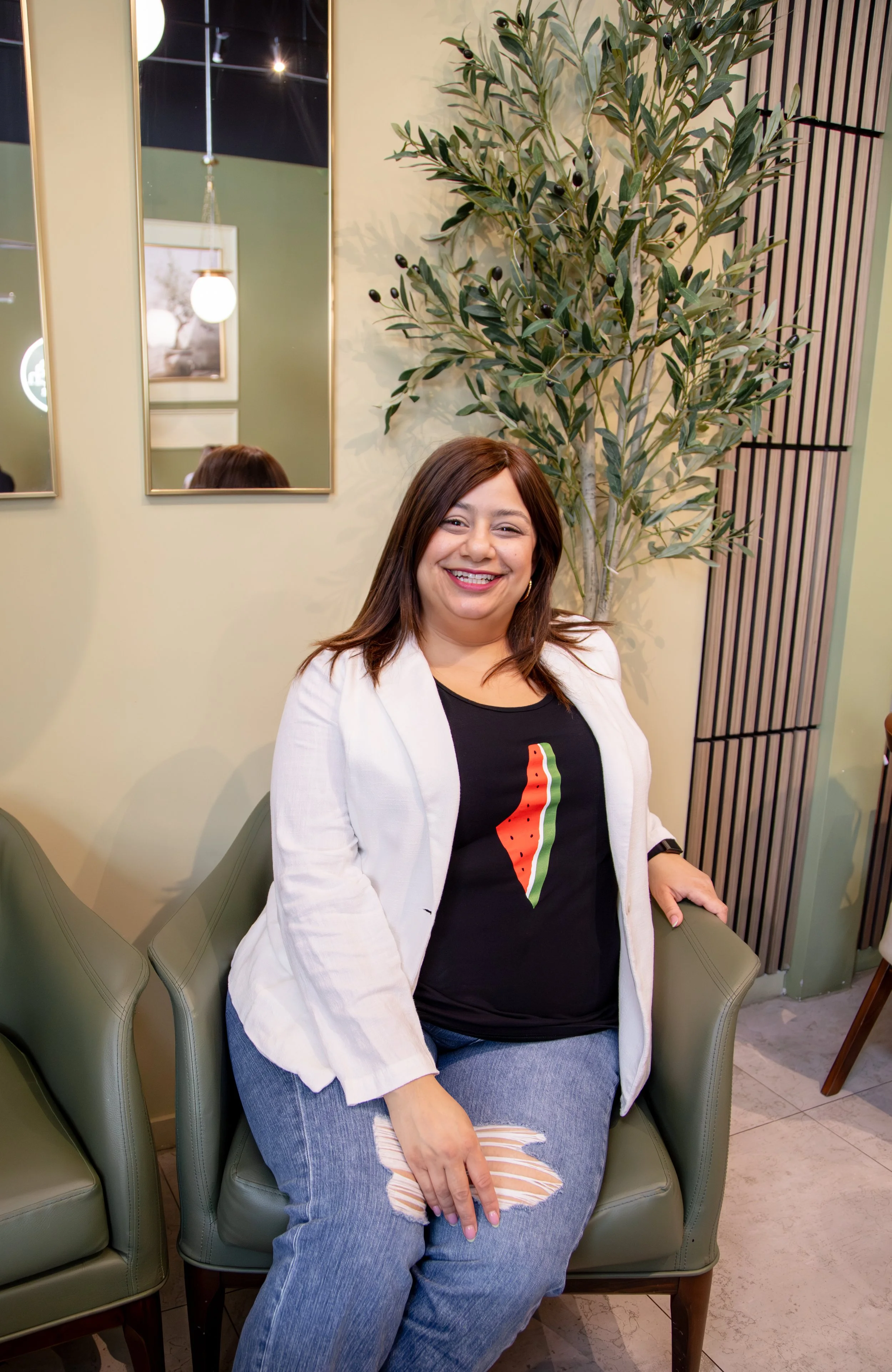 A woman with brown hair smiling and sitting in a green armchair in a modern interior with mirrors, wall art, a large plant, and wood panel accents.