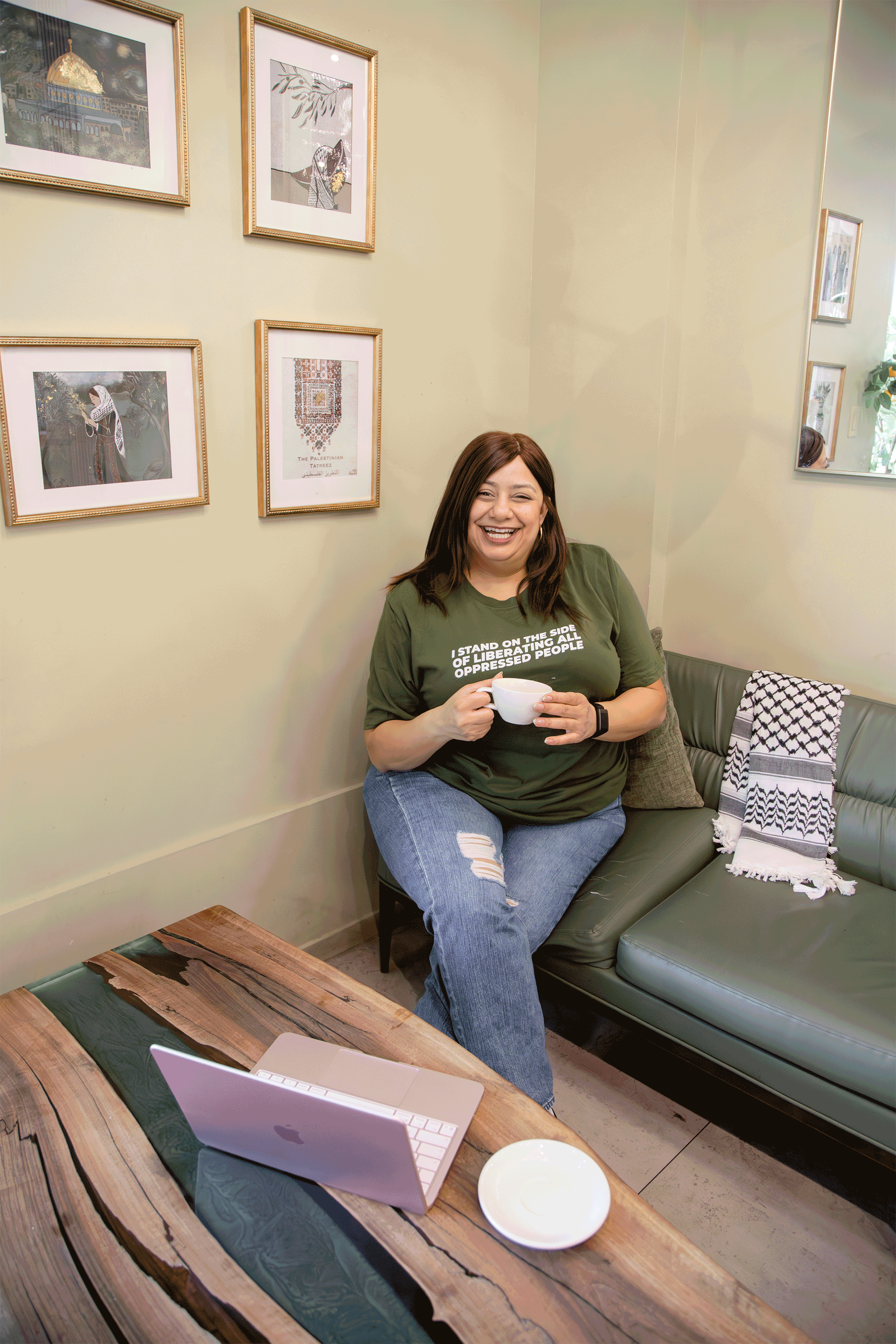 A woman with long brown hair, wearing a green T-shirt and ripped jeans, smiling and holding a white coffee cup while sitting on a green leather couch in a room decorated with framed artwork on the walls, a wooden coffee table, and a laptop on a wooden table.