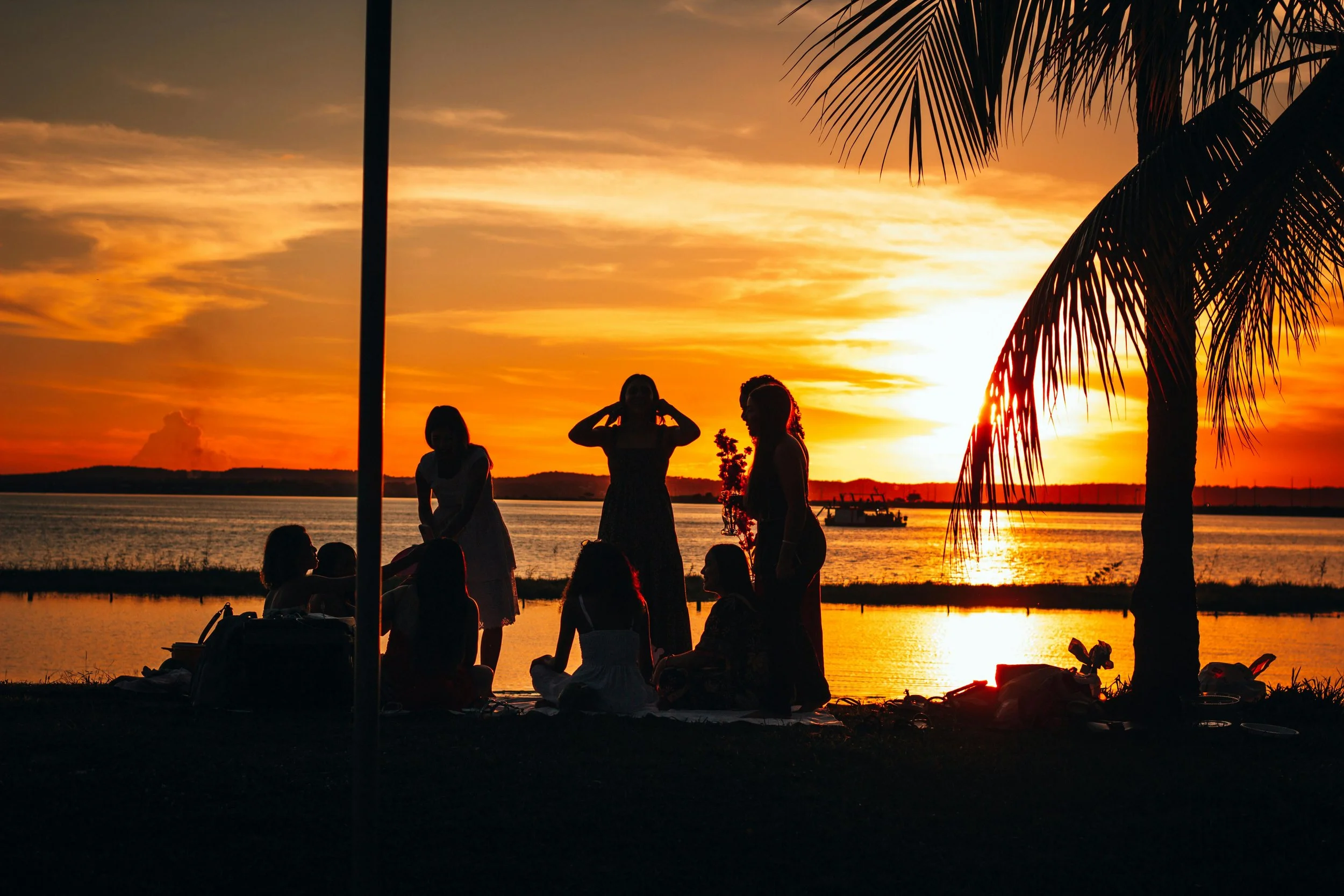 Silhouettes of people gathered outdoors during sunset near a body of water, with palm trees and a boat in the background.