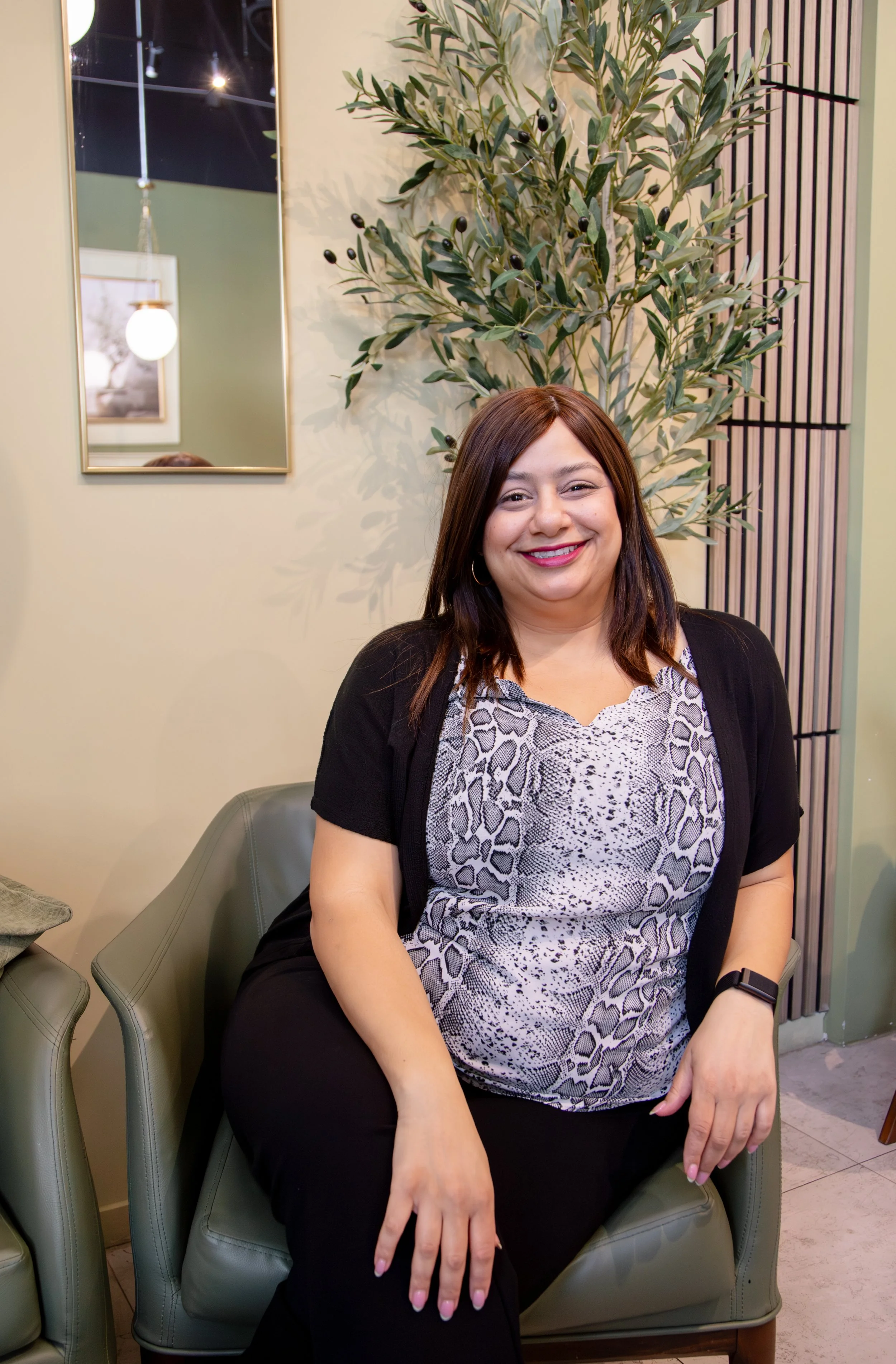 A woman with long brown hair, wearing a patterned blouse and black cardigan, sitting on a green armchair. She is smiling and has a watch on her left wrist. Behind her, there is a tall green plant with dark berries and a mirror on the wall reflecting a light fixture and picture frame.