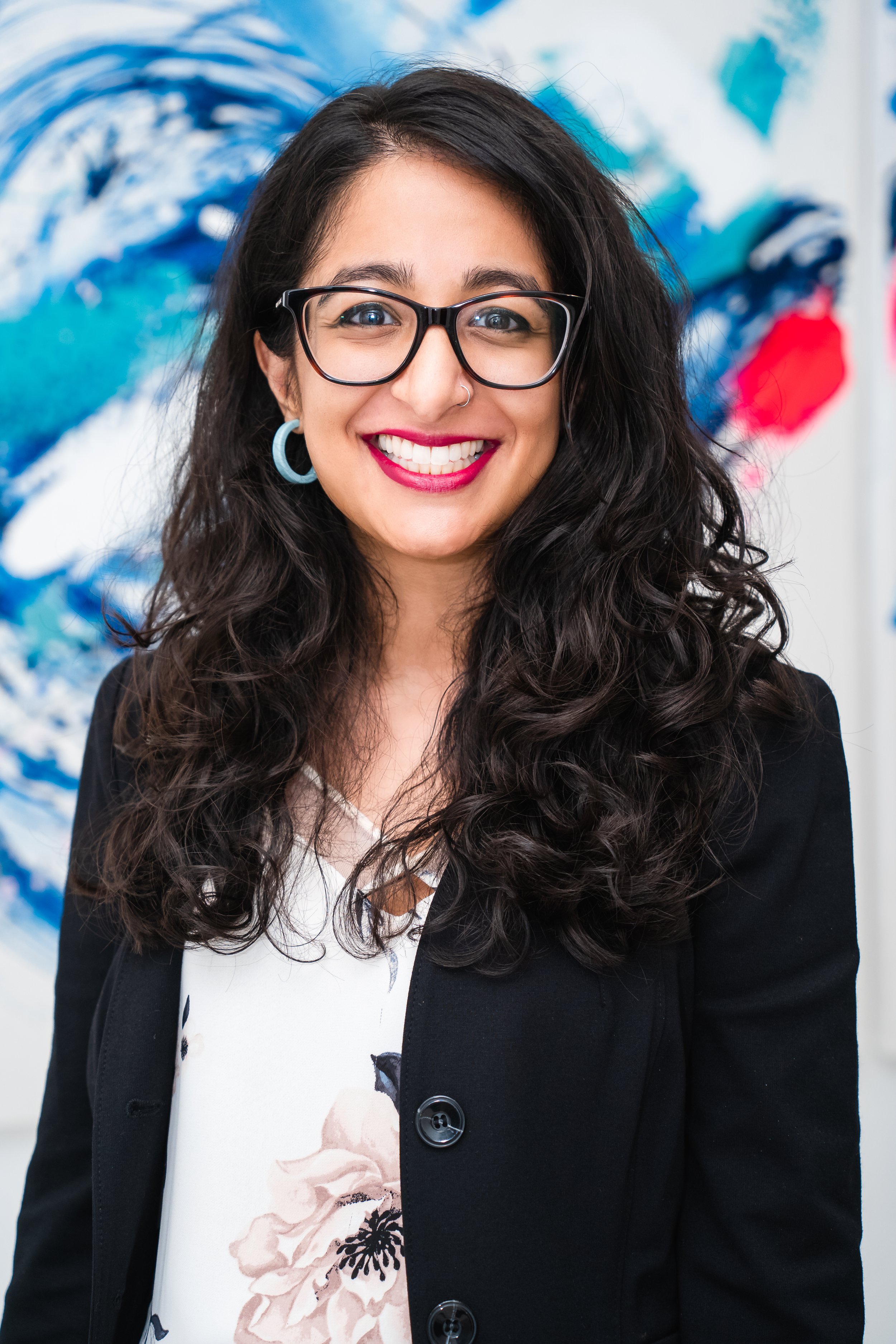 Woman with long curly dark hair, glasses, and red lipstick smiling, wearing a black blazer over a white floral top, standing in front of an abstract blue and red painting.