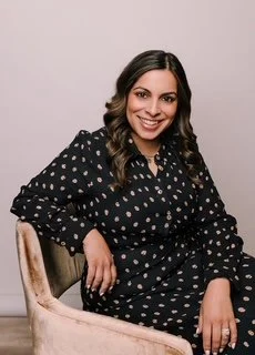 A woman with long dark hair wearing a black dress with white polka dots, sitting on a chair and smiling at the camera.