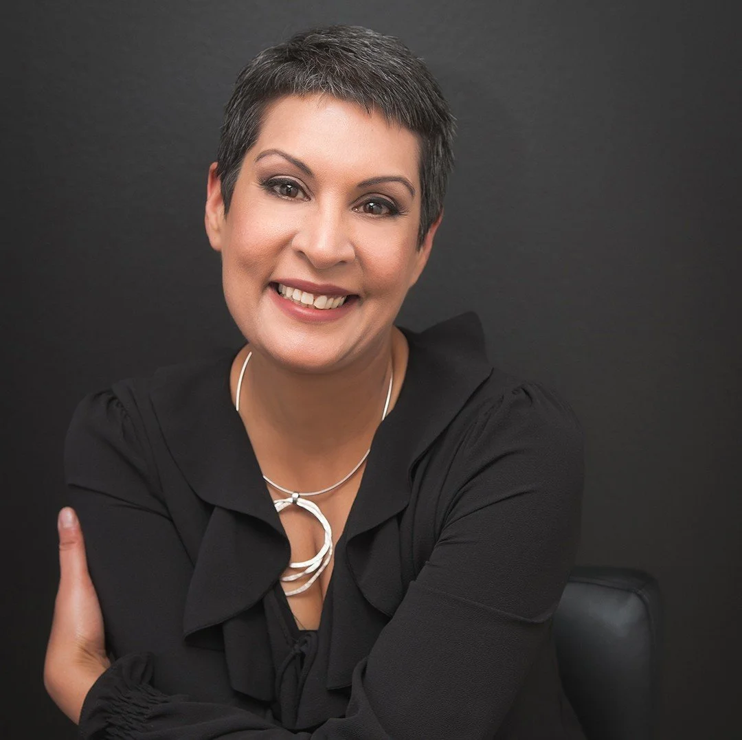 A woman with short dark hair smiling at the camera against a black background, wearing a black blouse and a silver necklace.