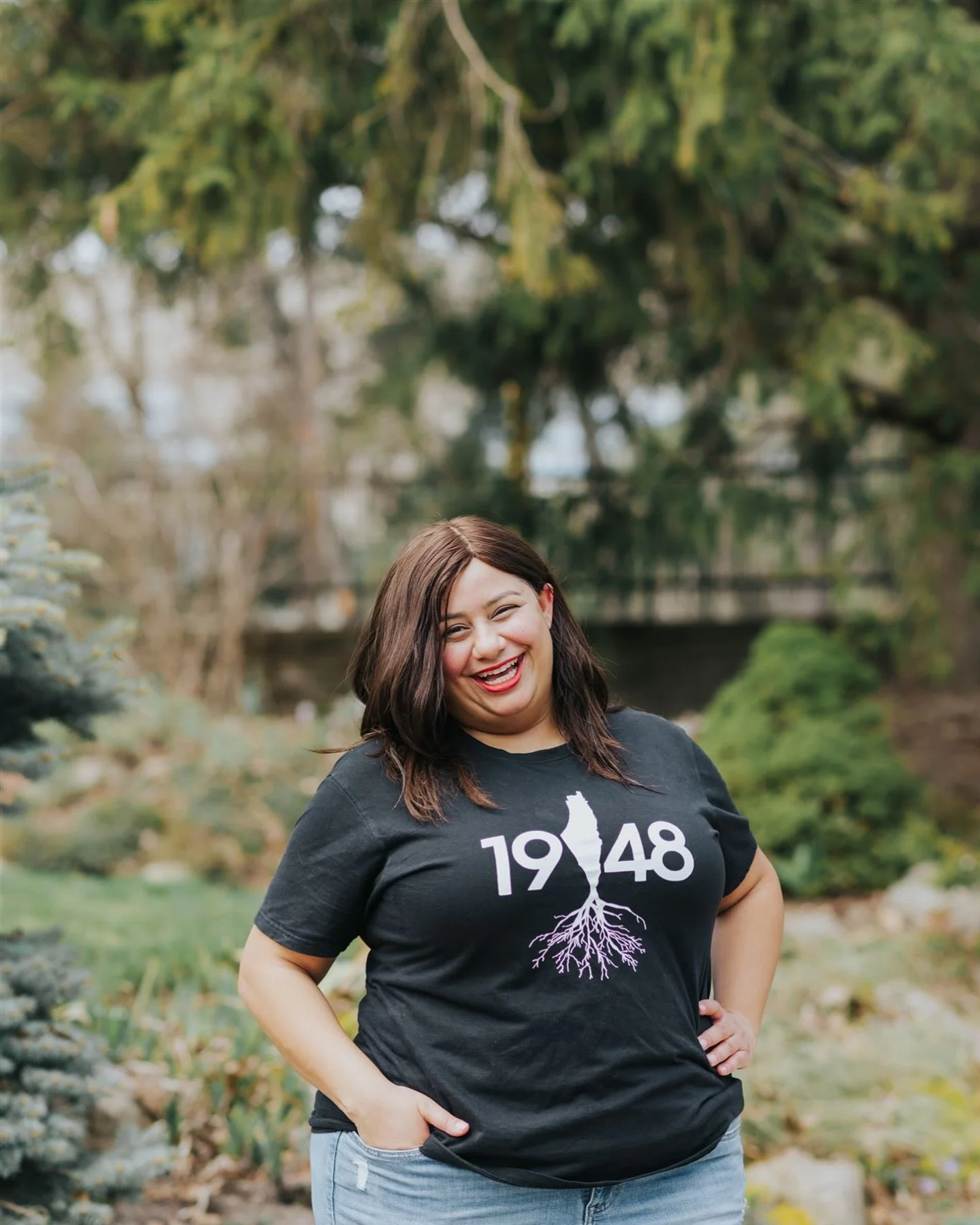 A woman with dark brown hair smiling outdoors in front of trees and a stream, wearing a black T-shirt with a white graphic of a plant in the form of Palestine and the year 1948.