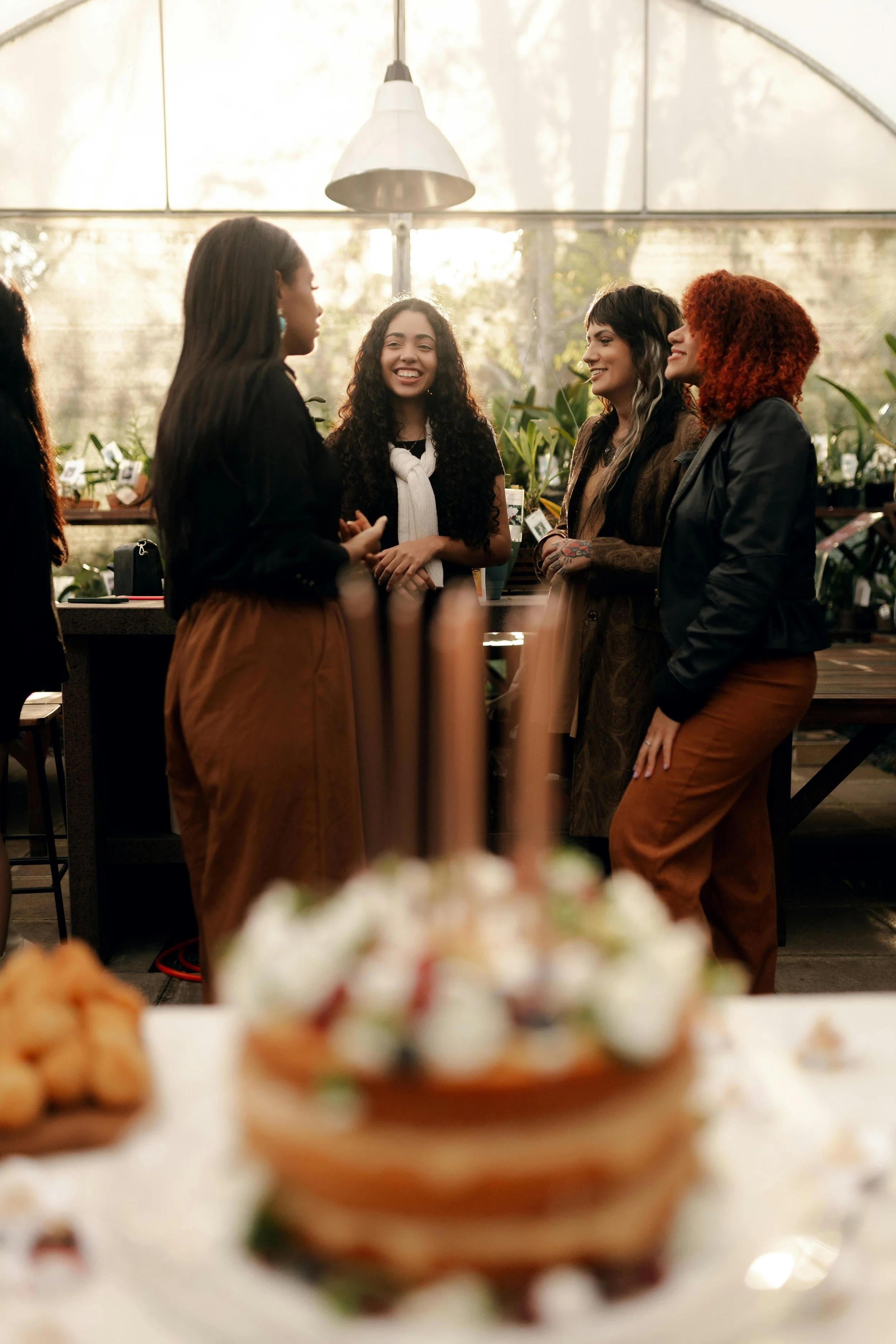 Four women conversing and smiling in a greenhouse with plants in the background, with blurred trays of food in the foreground.