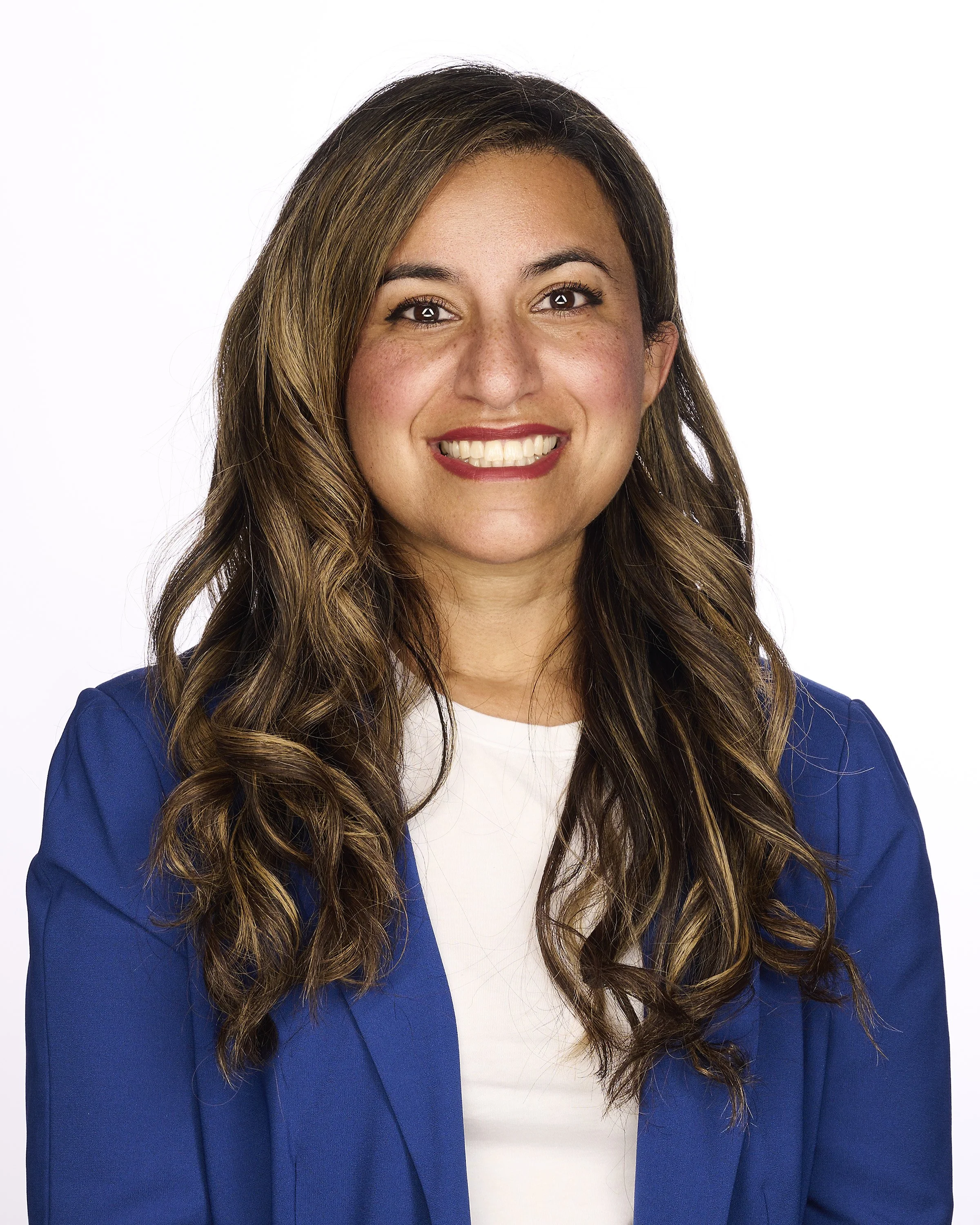A smiling woman with long, wavy brown hair and light skin, wearing a blue blazer over a white top, against a plain white background.