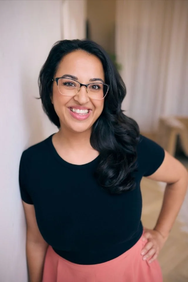A smiling woman with glasses and dark hair, wearing a black top and pink pants, standing indoors with her hand on her hip.