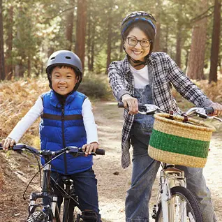 A woman and a boy riding bicycles on a wooded trail, both smiling, with the woman pushing the bike and carrying a basket in a forest setting.