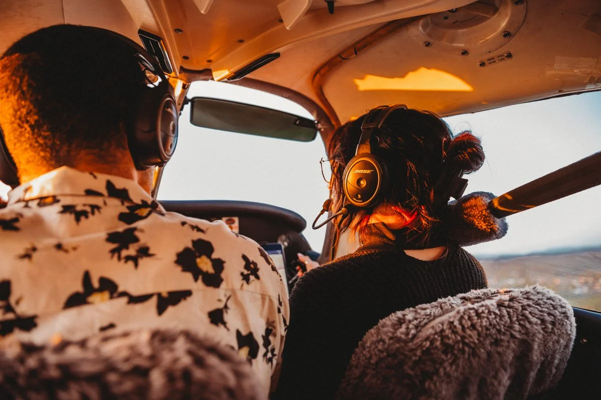 A young woman with dark hair, glasses, and a headset sitting in the cockpit of a small airplane during sunset, looking at a tablet.