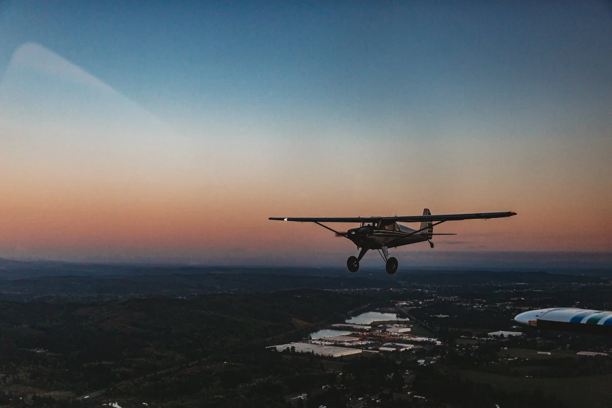 A small airplane flying over a landscape during sunset or sunrise, with bodies of water and forests visible below.