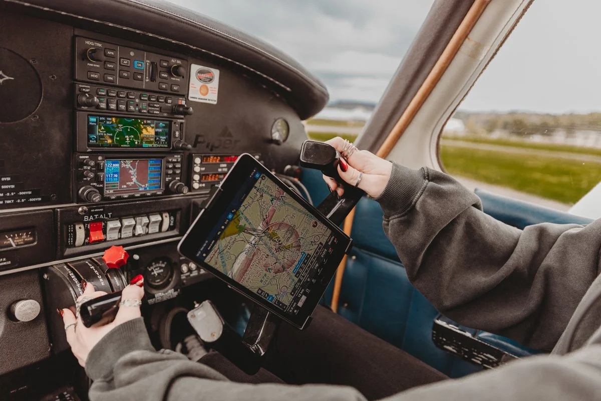 Pilot's hands in a cockpit, holding a tablet displaying a navigation map, with various airplane instruments on the panel.