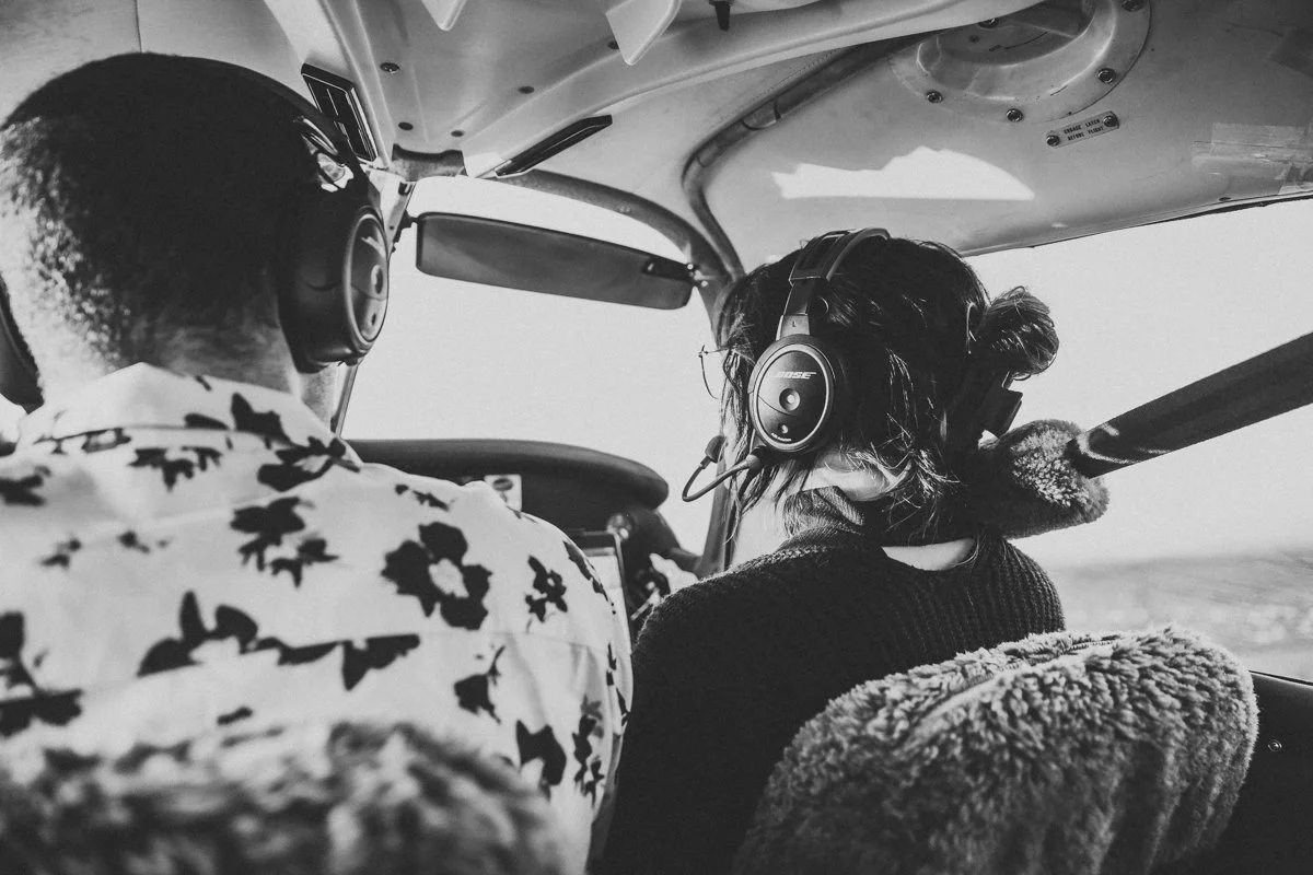 A pilot and passenger inside a small aircraft cockpit, both wearing headphones, with the pilot operating the controls and the passenger looking out the window.