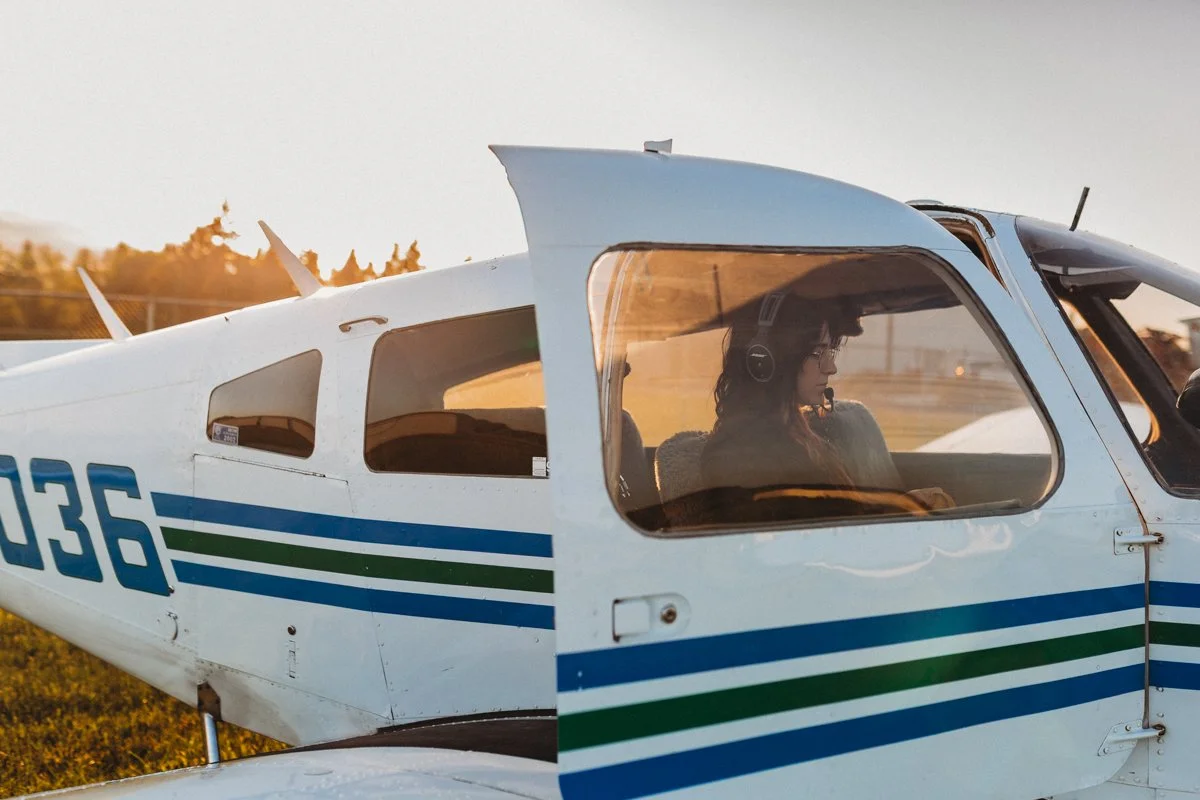 A woman wearing headphones sitting inside a small white aircraft with blue and green stripes, parked outdoors during sunset.