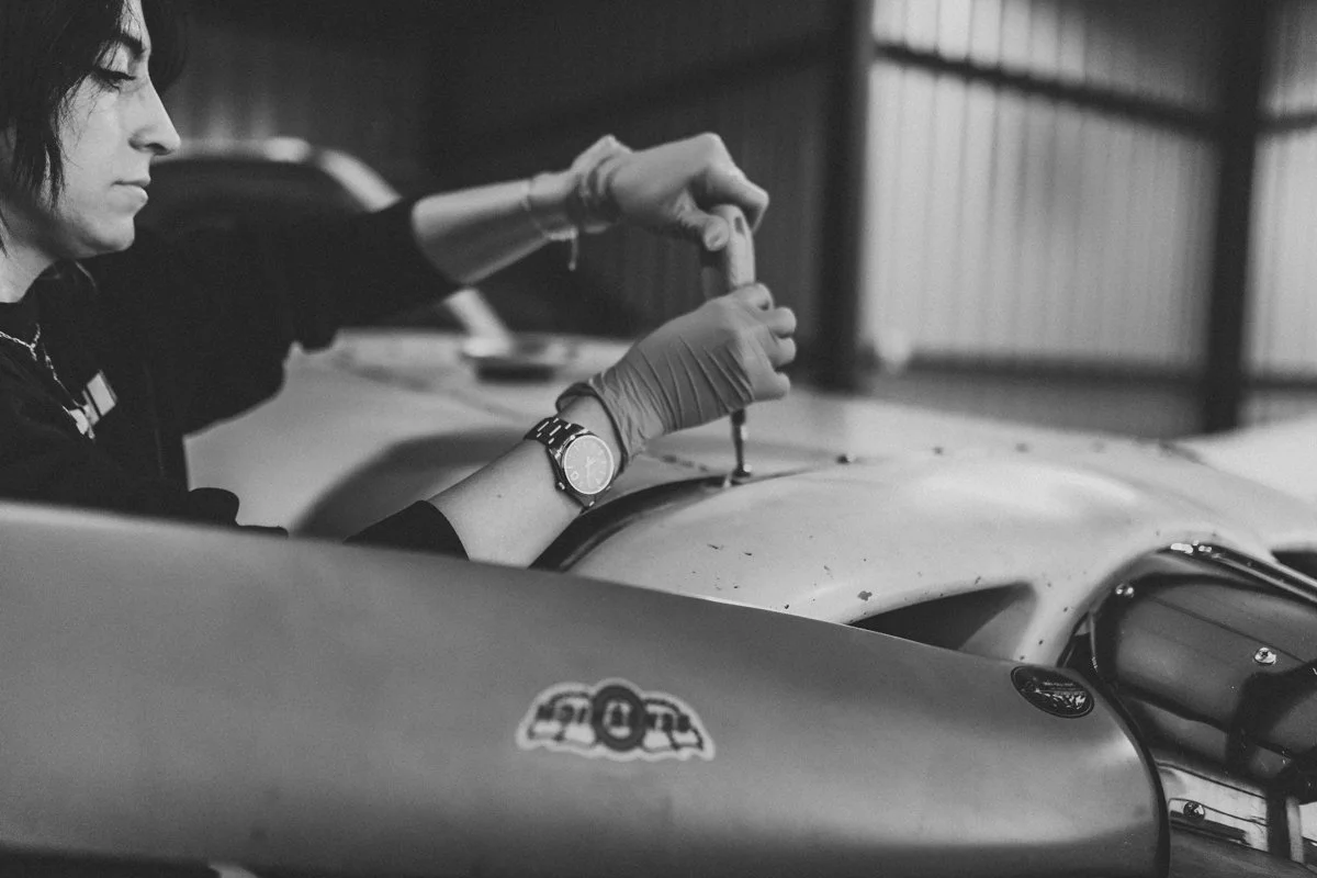 A woman wearing gloves and a wristwatch working on a vintage race car in a garage or workshop.