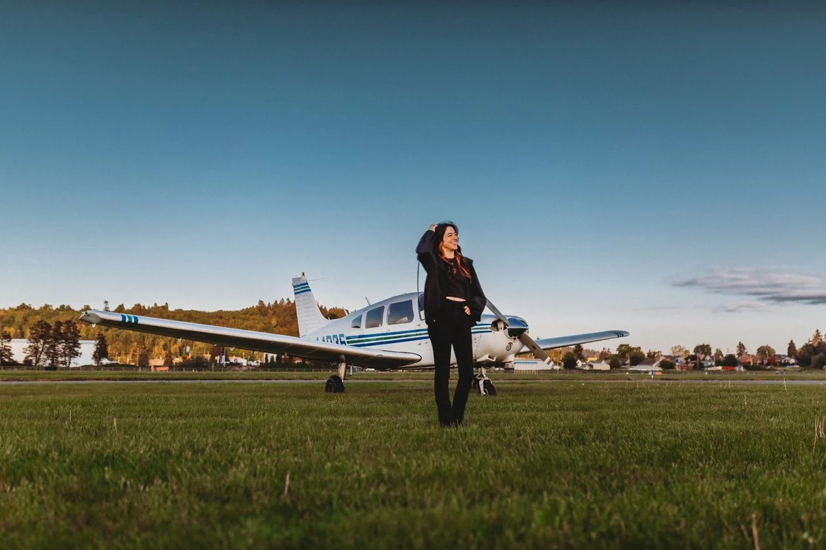 A woman standing on a grassy airfield near a small private airplane, with trees and houses in the background under a clear blue sky.