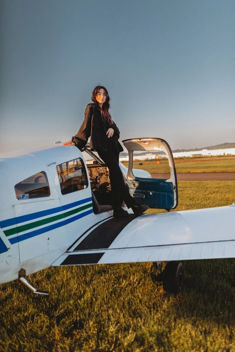 Woman sitting on the wing of a small airplane on a grassy field during sunset.