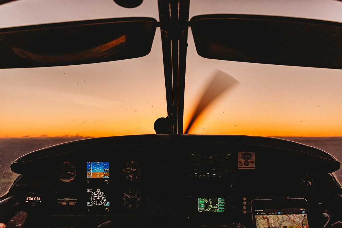 Inside an airplane cockpit at sunset, showing flight instruments, a GPS device, and a smartphone with a map.