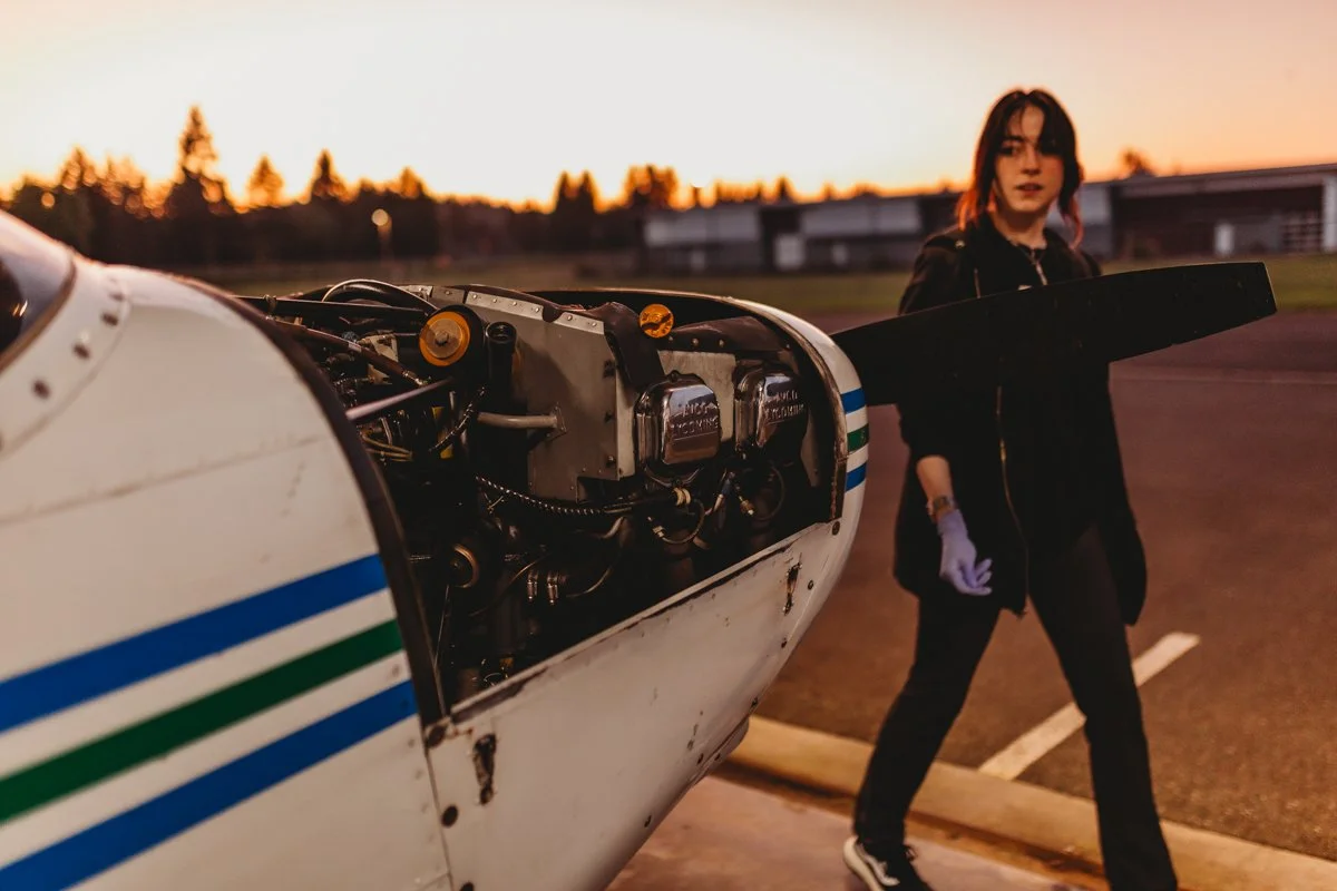 A woman with dark hair and a black jacket holding a wing of a small aircraft on a runway at sunset.