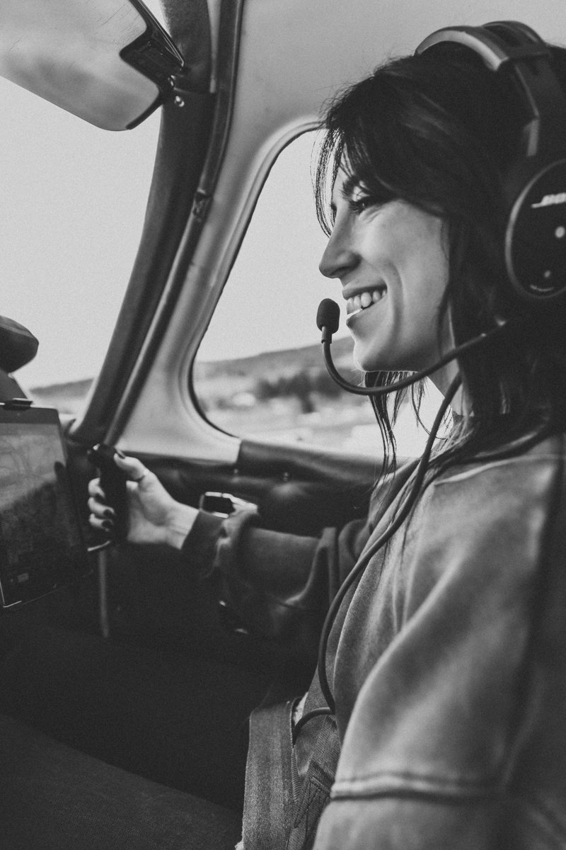 A woman smiling while flying a small aircraft, sitting in the cockpit with a headset on and holding the controls.