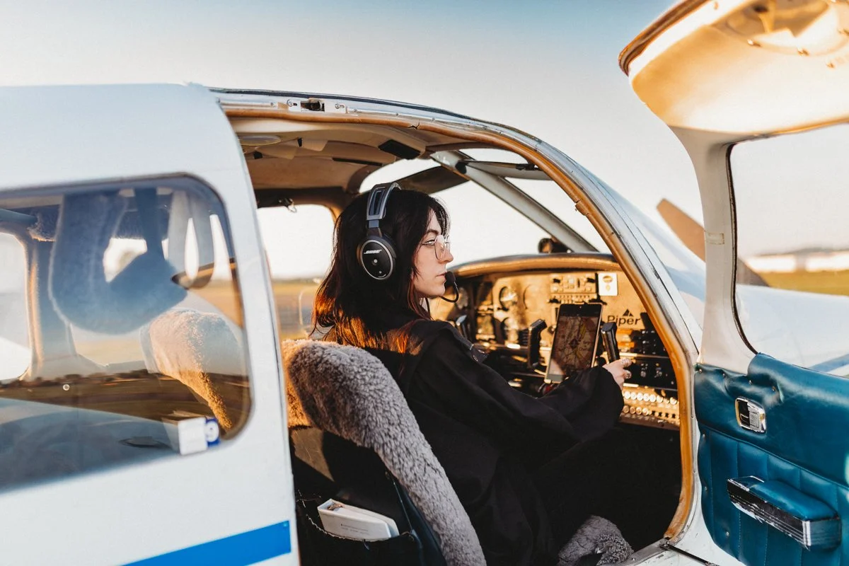 A woman with dark hair wearing glasses and a headset is sitting in the cockpit of a small airplane, holding a device and looking at the instrument panel during sunset.