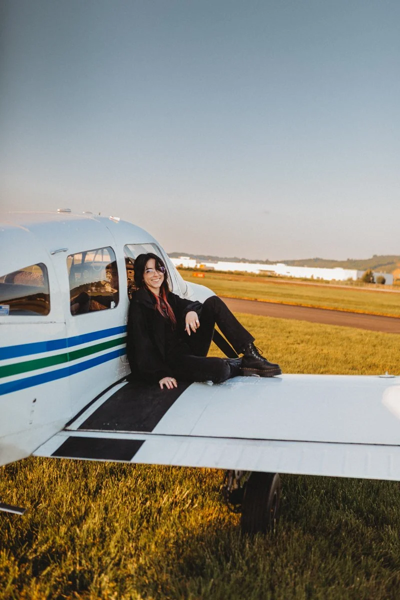 A woman sitting on the wing of a small airplane on a grassy airfield during sunset, smiling at the camera.