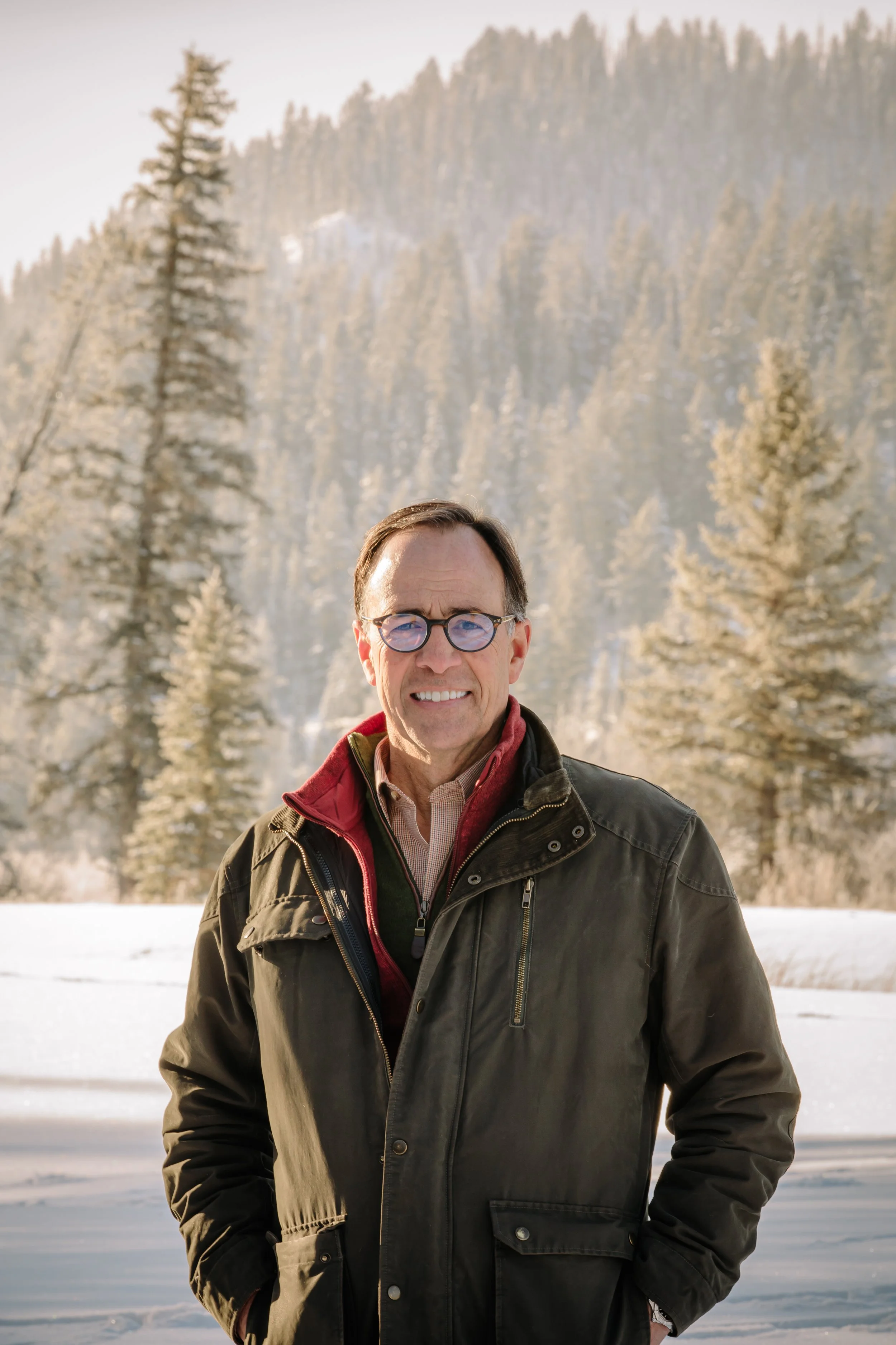 A middle-aged man with glasses smiling outdoors in a snowy landscape with tall evergreen trees and mountains in the background.