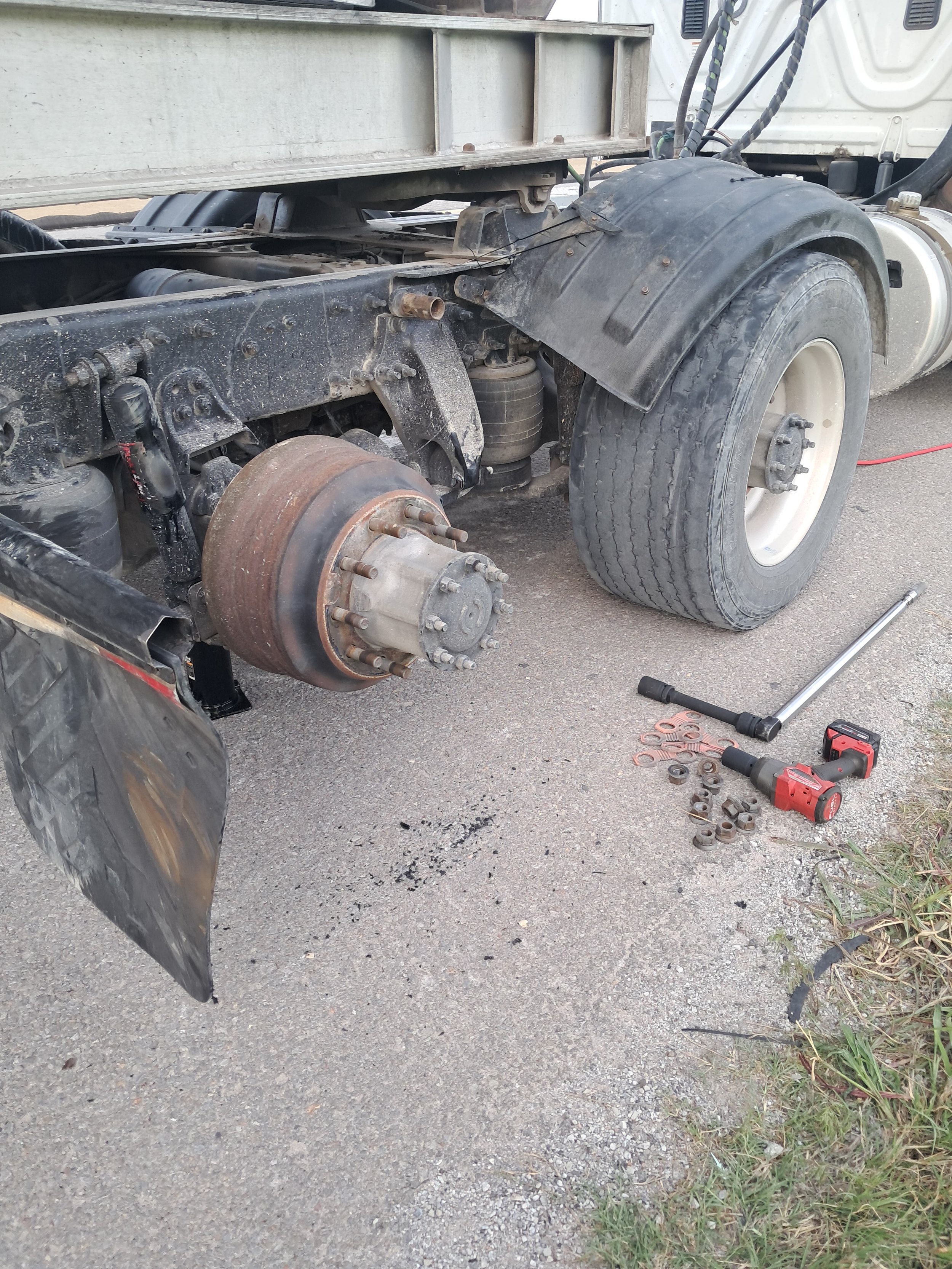 A close-up of a truck's rear wheel area with tire removed, exposing suspension and brake components. Tools and lug nuts are on the ground nearby, indicating maintenance work.