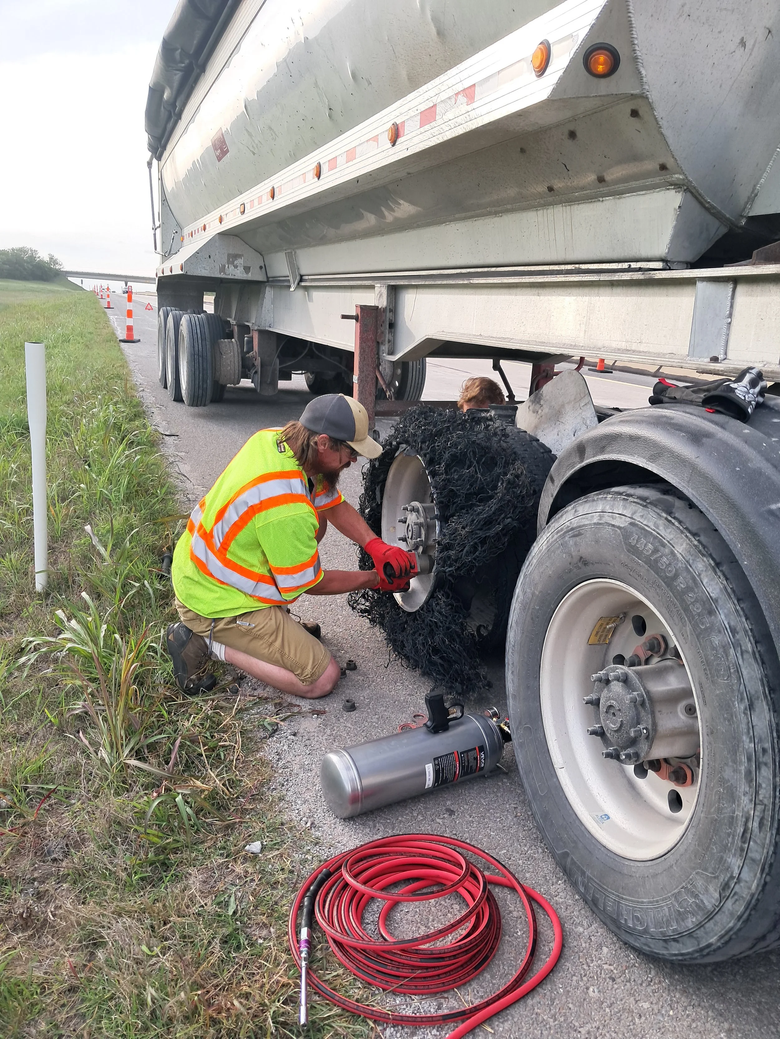 Roadside repair of a semi truck tire, with tools and equipment around, and traffic cones in the background.