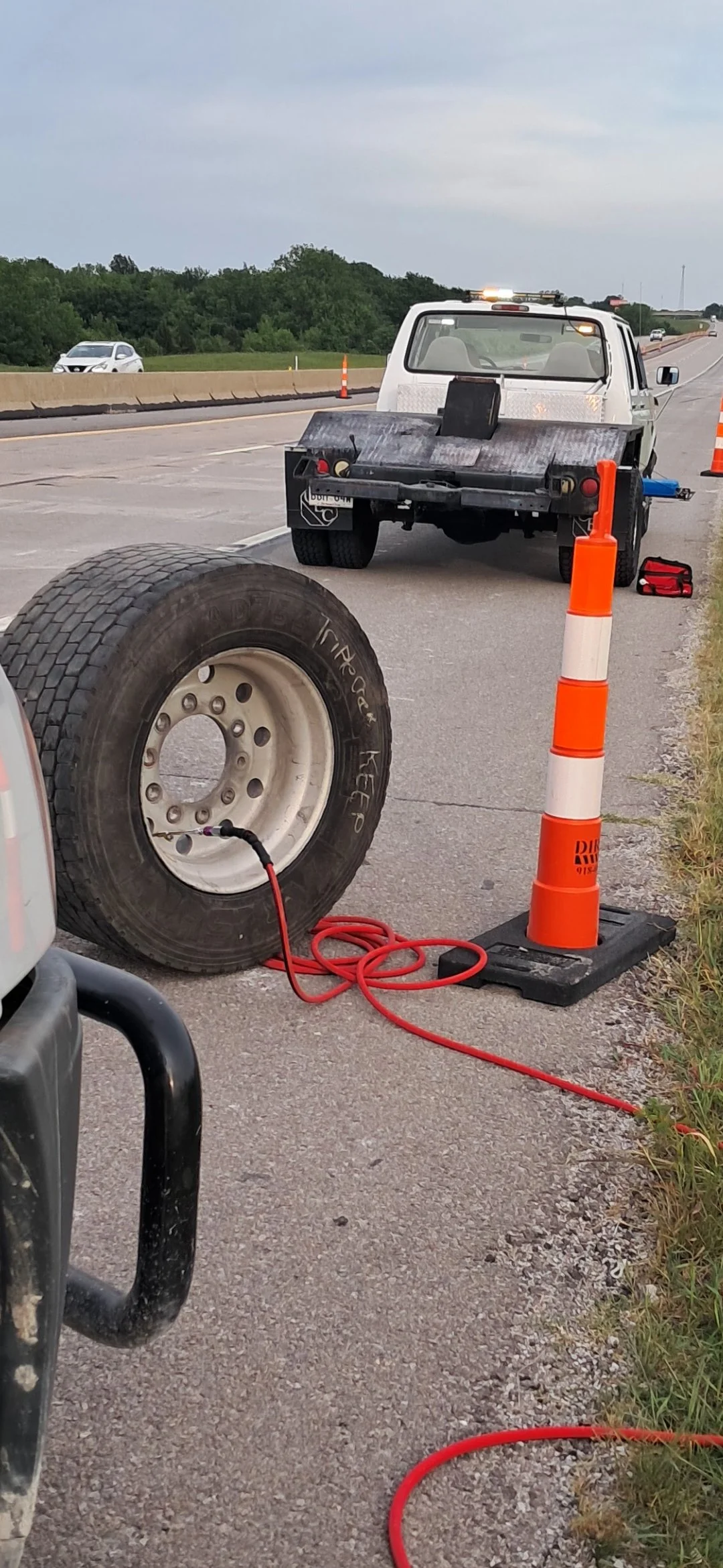 A roadside scene on a highway with traffic cones and a white roadside service truck parked on the side of the road. A super single semi tire is in the foreground with a red air hose connected filling up with air.