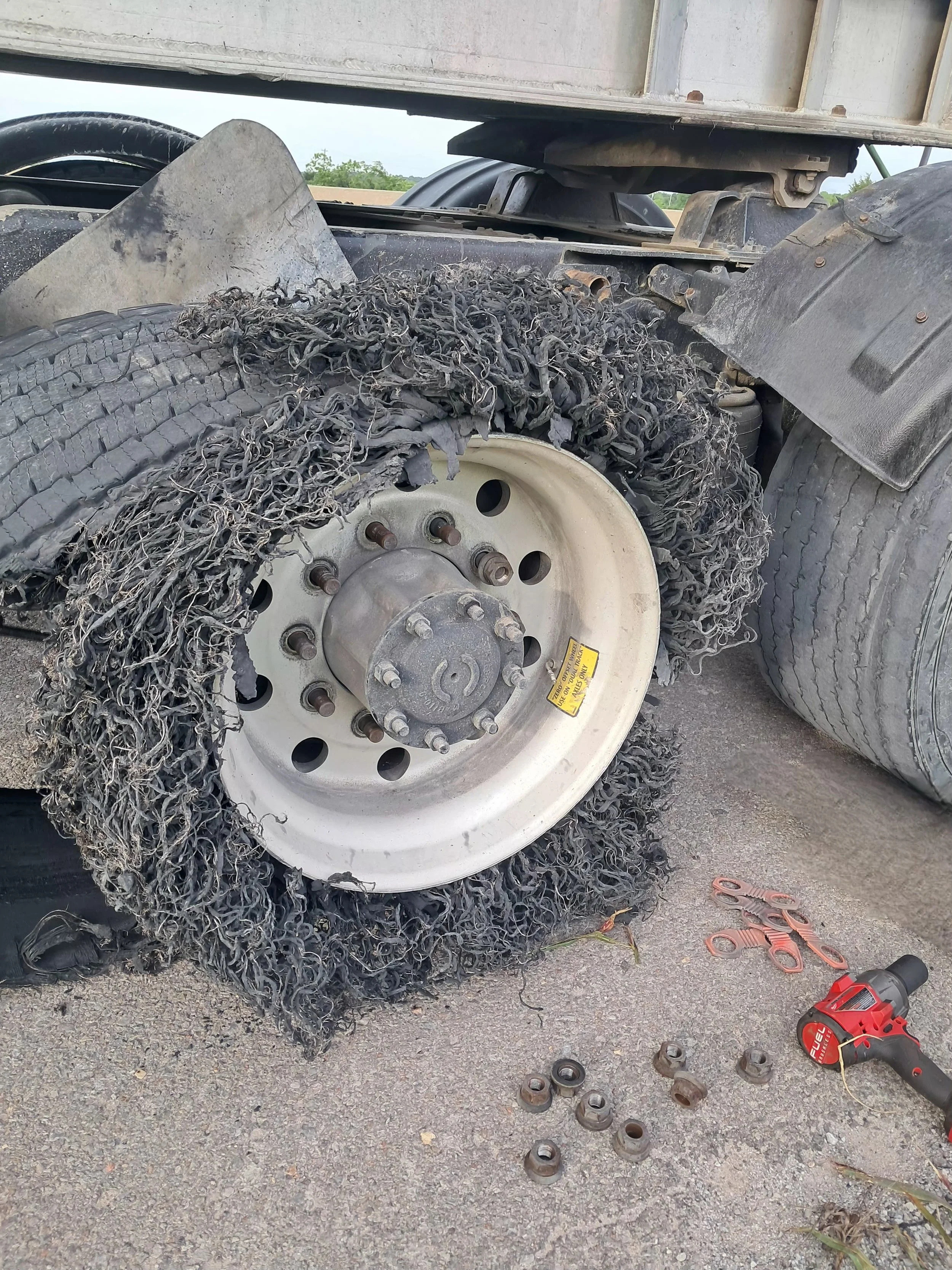 Close-up of a semi truck wheel with a blown super single tire, surrounded by tools and loose wheel nuts on the ground in preparation for a tire change.