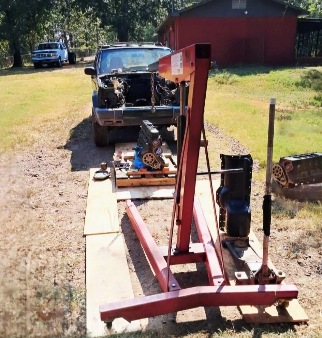 An outdoor scene with a Jeep Cherokee in a yard undergoing an engine swap, with an engine hoist in the foreground and an engine that was delivered on a pallet ready to swap.