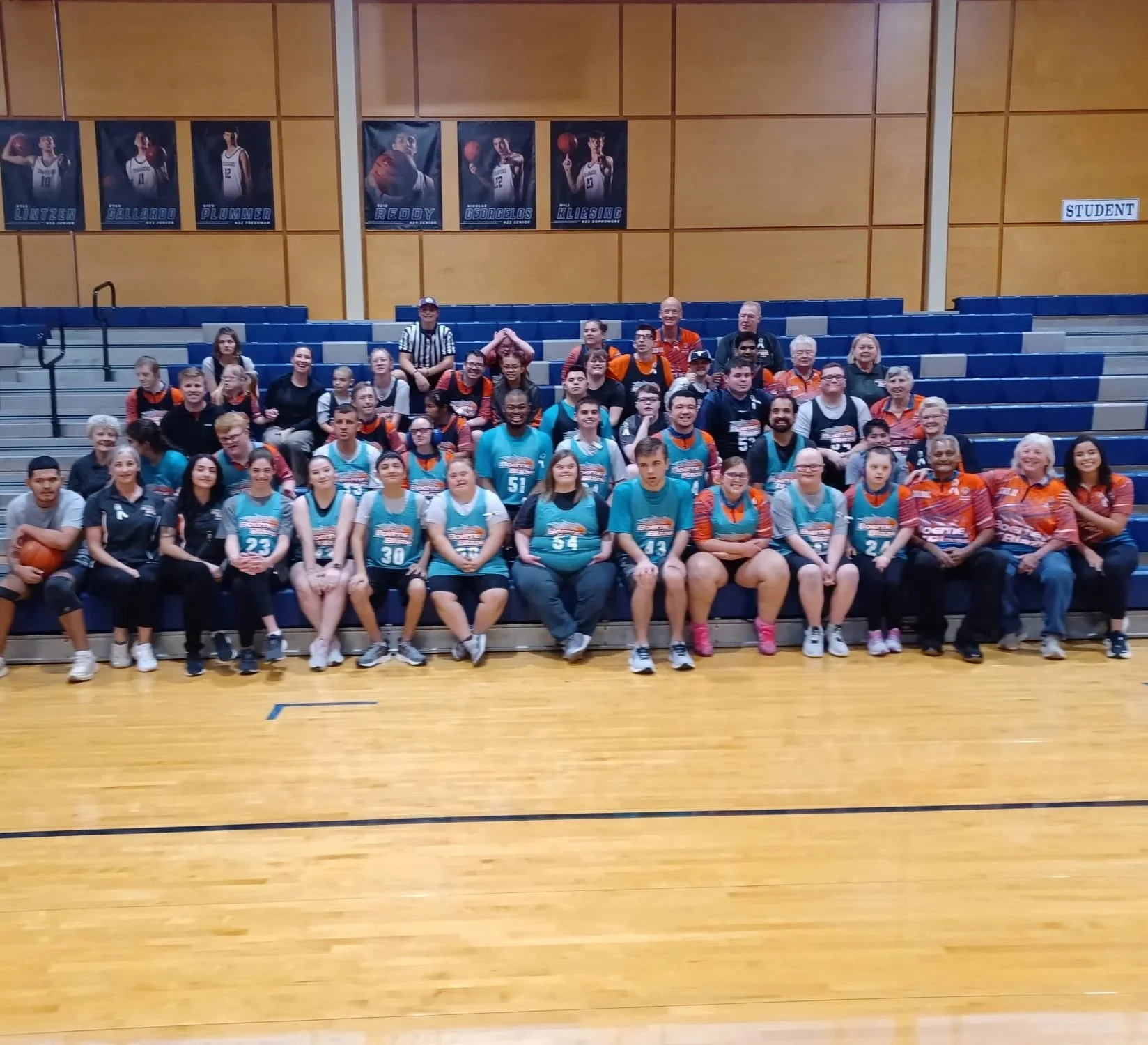 A large group of people, including children and adults, posing for a photo in an indoor gymnasium. The group includes basketball players in turquoise and orange jerseys, and others in casual and team attire. They are seated and standing in front of blue bleachers, with posters of basketball players on the wall and a sign labeled