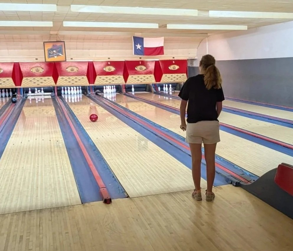 A woman standing at the edge of a bowling lane, watching a red bowling ball roll down the lane towards the pins. In the background, there is a Texas state flag and bowling lanes with pins set up.