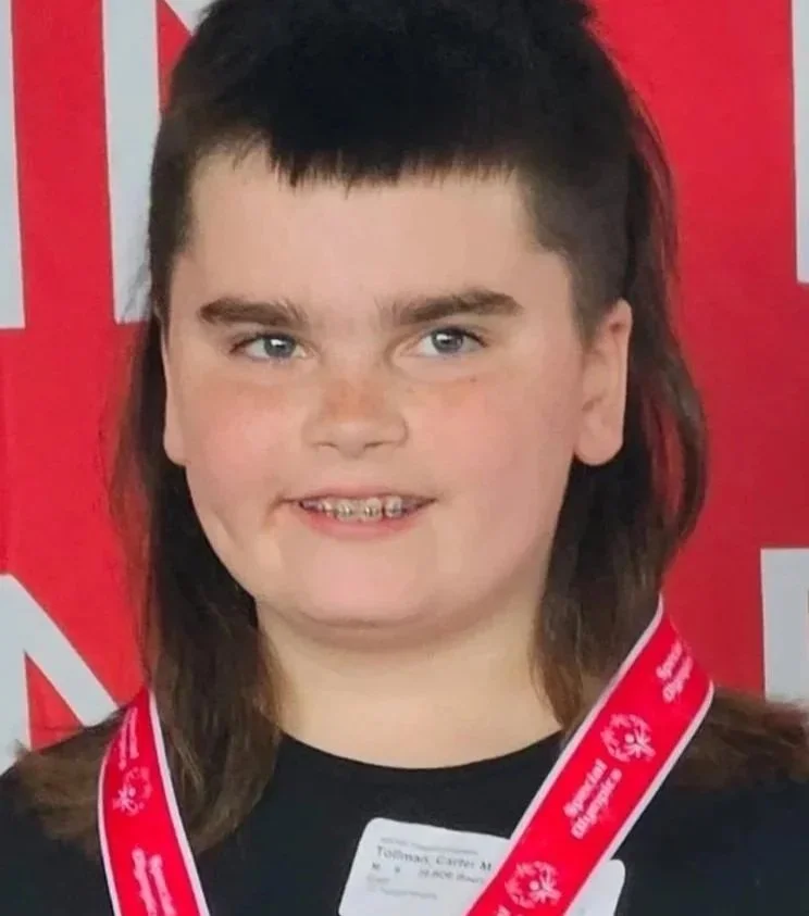 A young girl with dark hair and light skin, wearing a red ribbon with white snowflakes and text, standing in front of a red background.