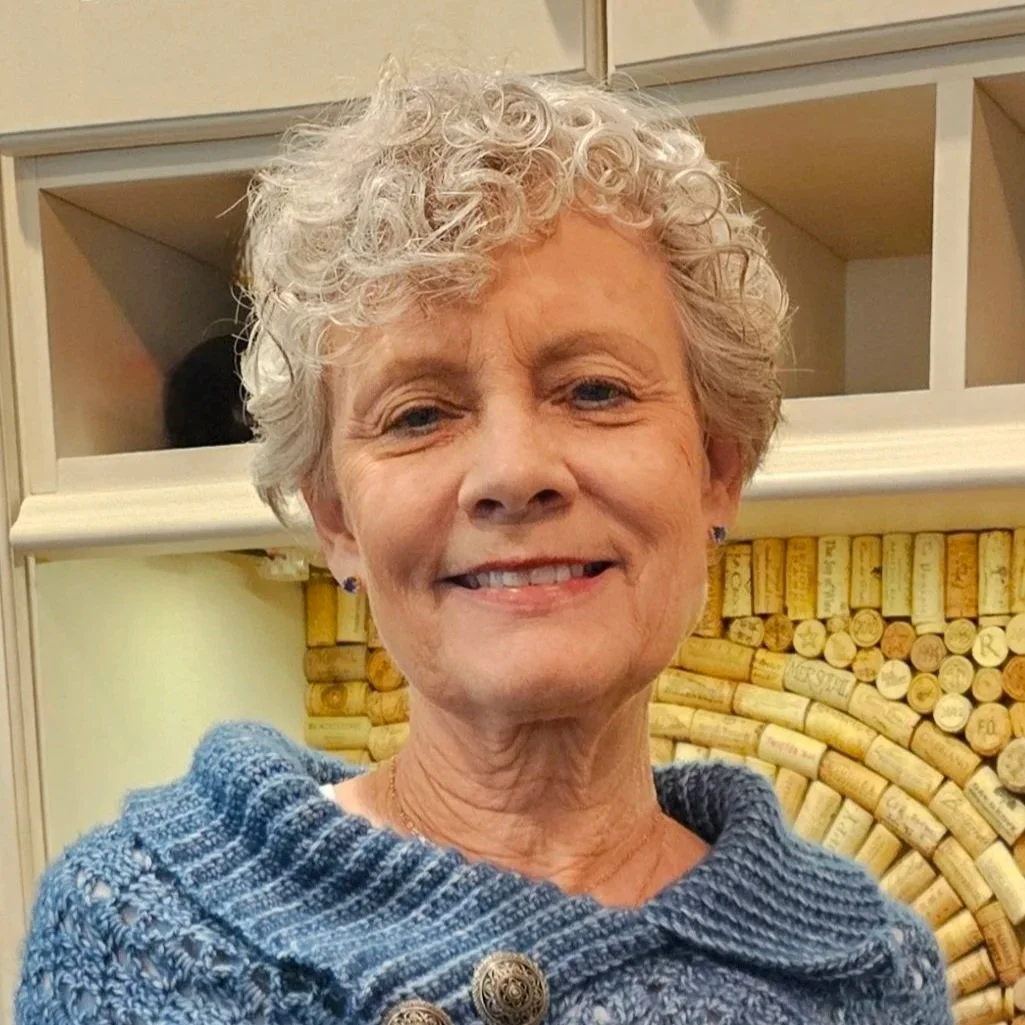 A smiling older woman with curly gray hair wearing a blue knitted sweater with decorative buttons.