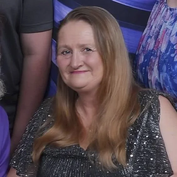 A woman with long, light brown hair smiling, wearing a black, sparkly top, in a group photo with other people.